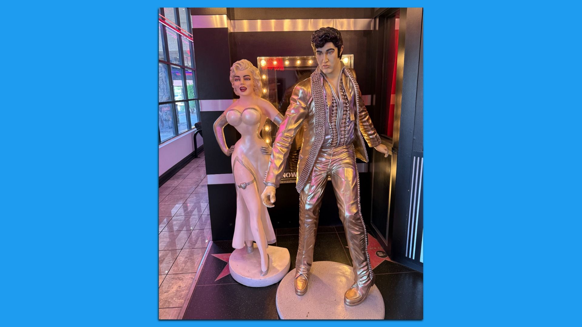 Life-size statues of Marilyn Monroe in a pink strapless gown and Elvis Presley in a shiny metallic gold suit, posed beside a lit vanity mirror in a display area.