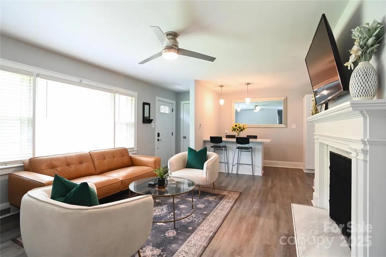 Bright living room with large window, tan leather couch, two cream armchairs with green pillows, round glass coffee table on patterned rug, ceiling fan, white fireplace, TV, and dining counter with pendant lights.