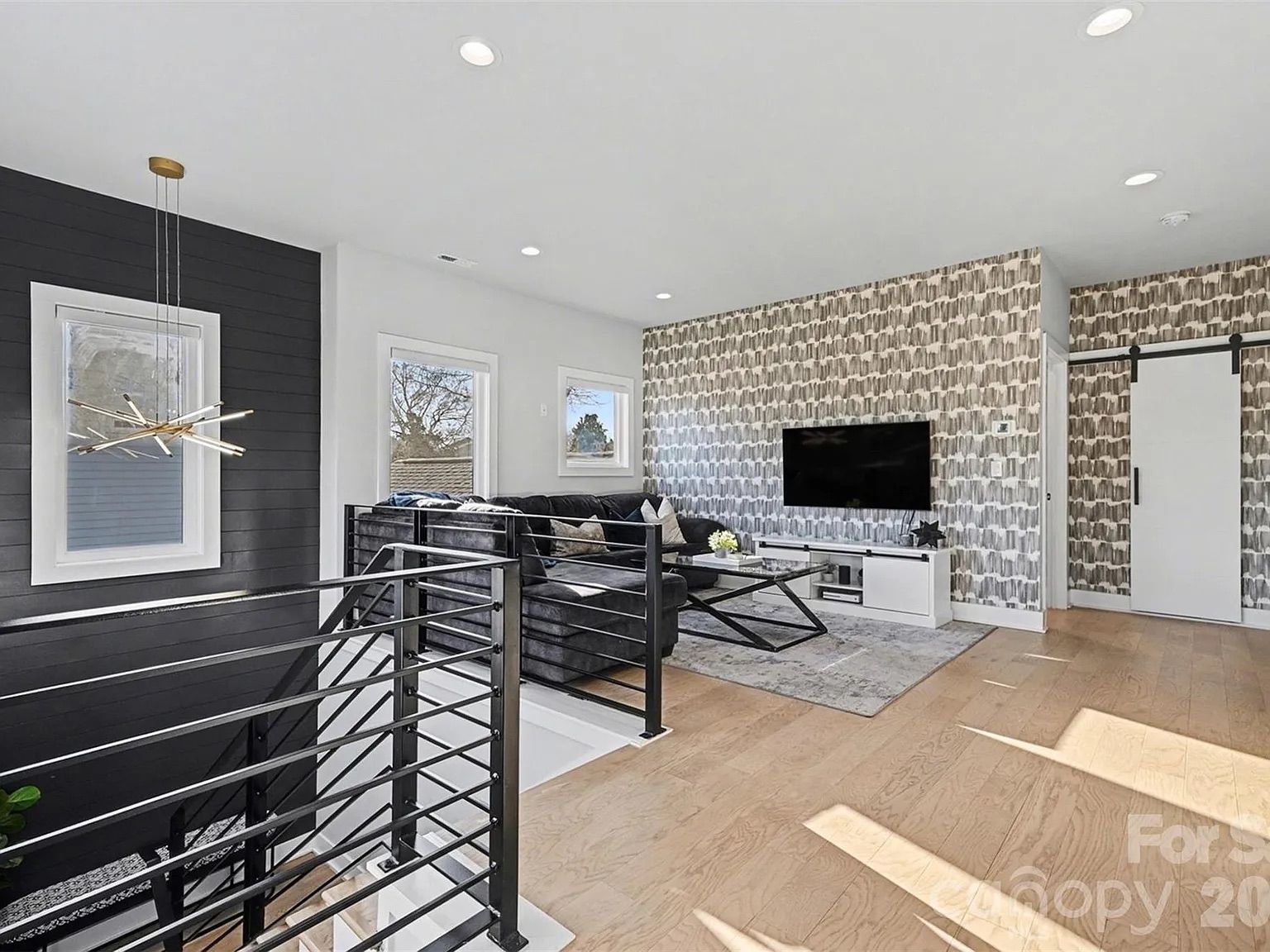 Bright modern living room with light wood floors, a black accent wall and a gold chandelier hanging near the window. A patterned wallpaper wall holds a mounted TV above a white console; a gray sectional sofa and glass coffee table sit on a light rug.