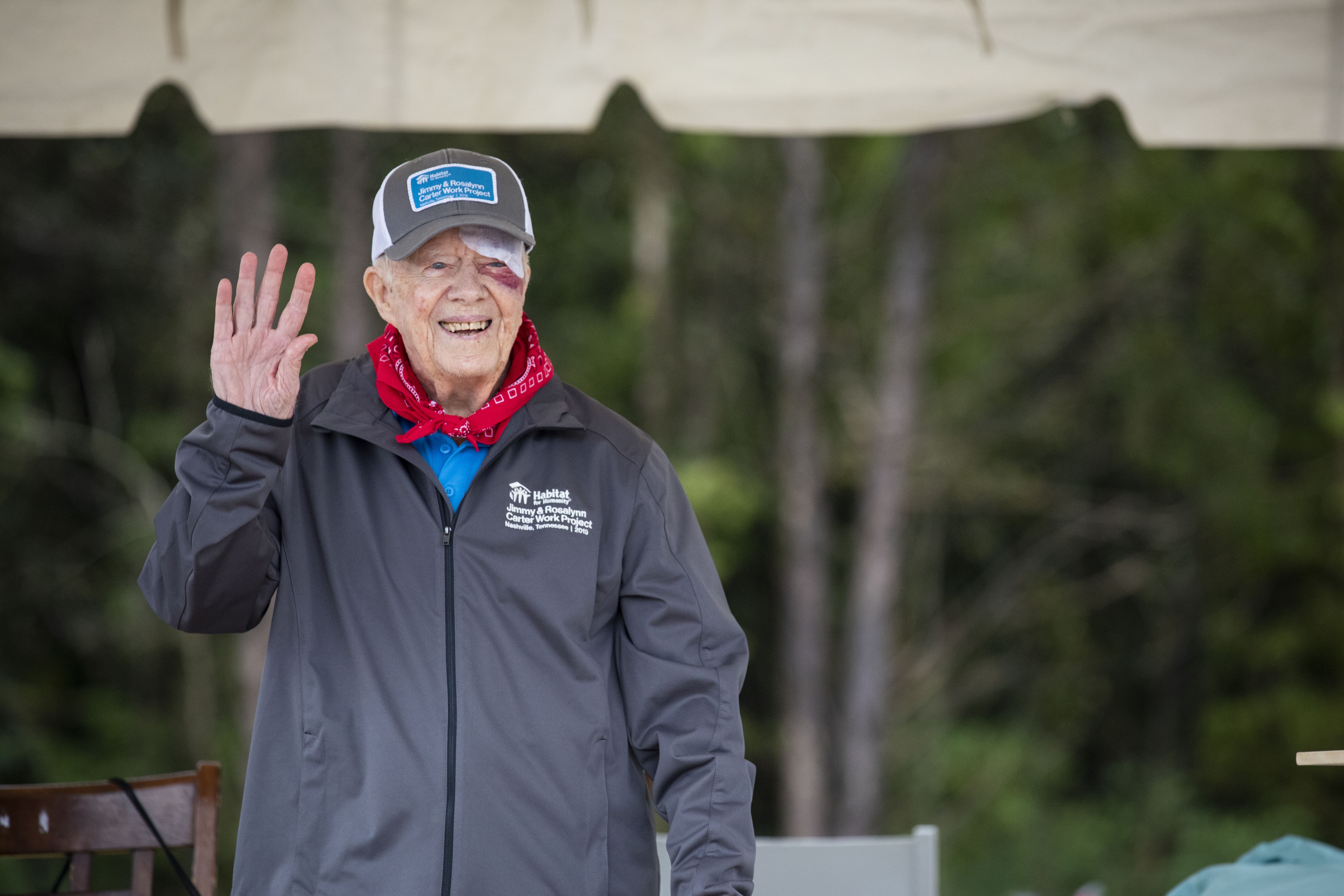 Jimmy Carter, 95 years old, waves to people while wearing a gauze patch over a black eye, with a red bandana around his neck and a jacket