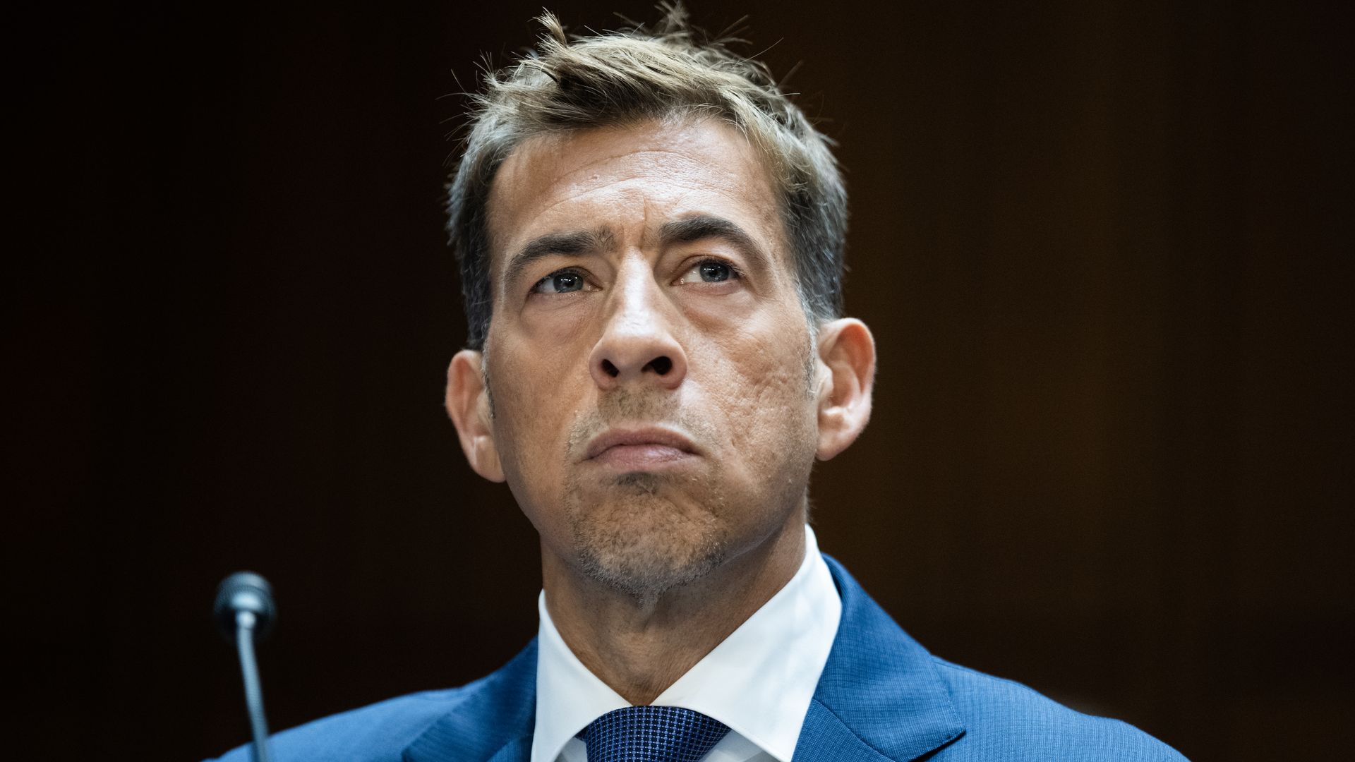 Close-up of a man in a bright blue suit and matching tie at a podium with a microphone, looking off to the side; dark, out-of-focus background.