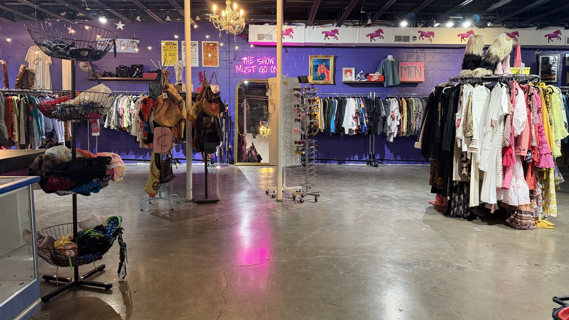 Interior of a vintage thrift store: purple walls, racks full of colorful dresses and jackets, bags on stands, a chandelier, neon pink sign says "THE SHOW MUST GO ON", and a "MEN" section in back.