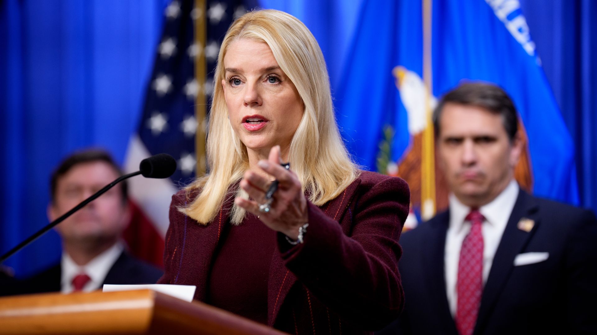 A bleached-blonde, long-haired Attorney General Pam Bondi, wearing a velvet maroon jacket and top, gestures with her left hand as she speaks to a reporter (off camera) at a podium in front of a blue curtain with the DOJ and US flags as two dark-haired, white, suited men look on.