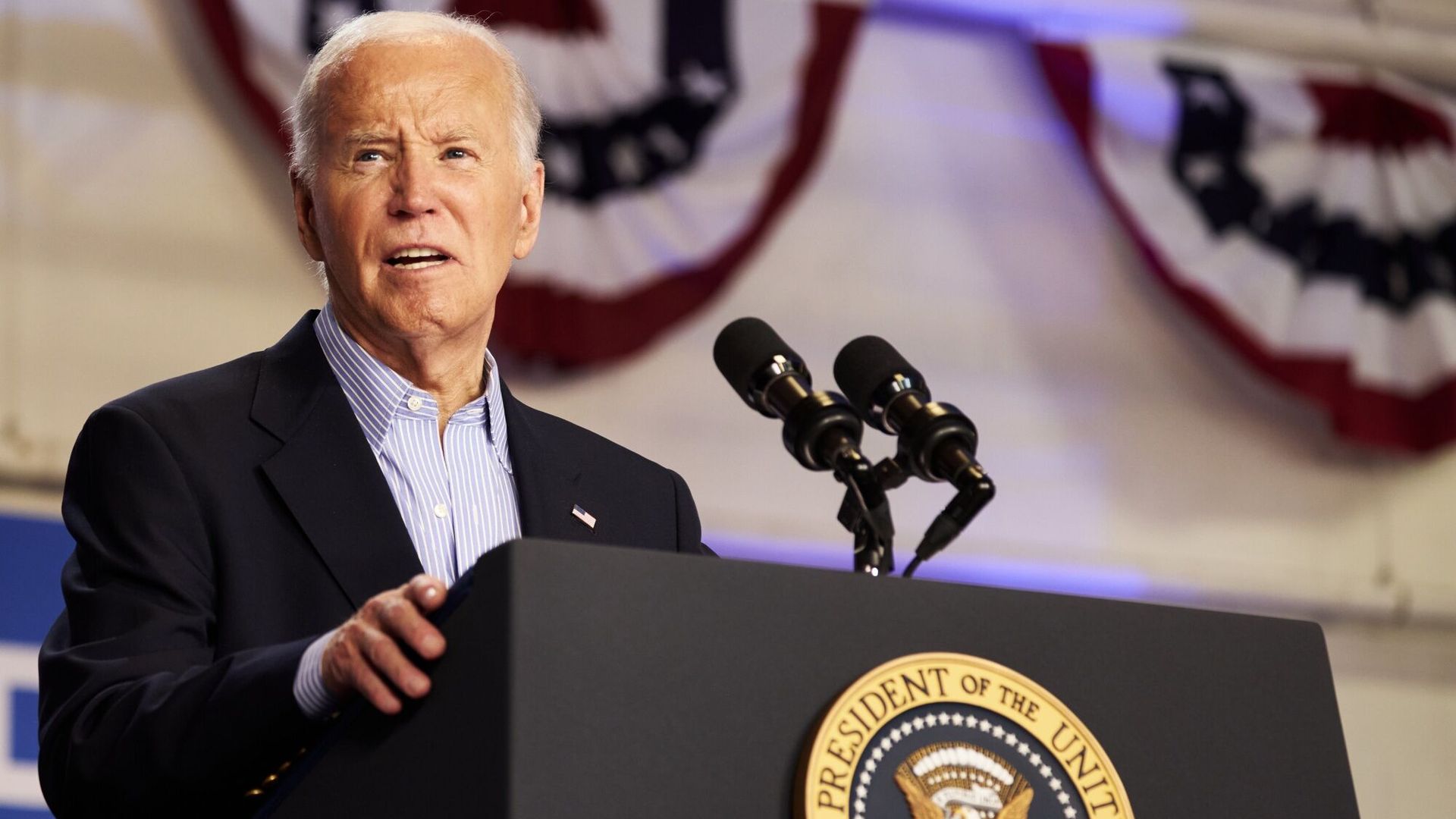 President Biden, wearing a dark gray suit and gripping a podium in front of a white wall with Valley Forge flags.