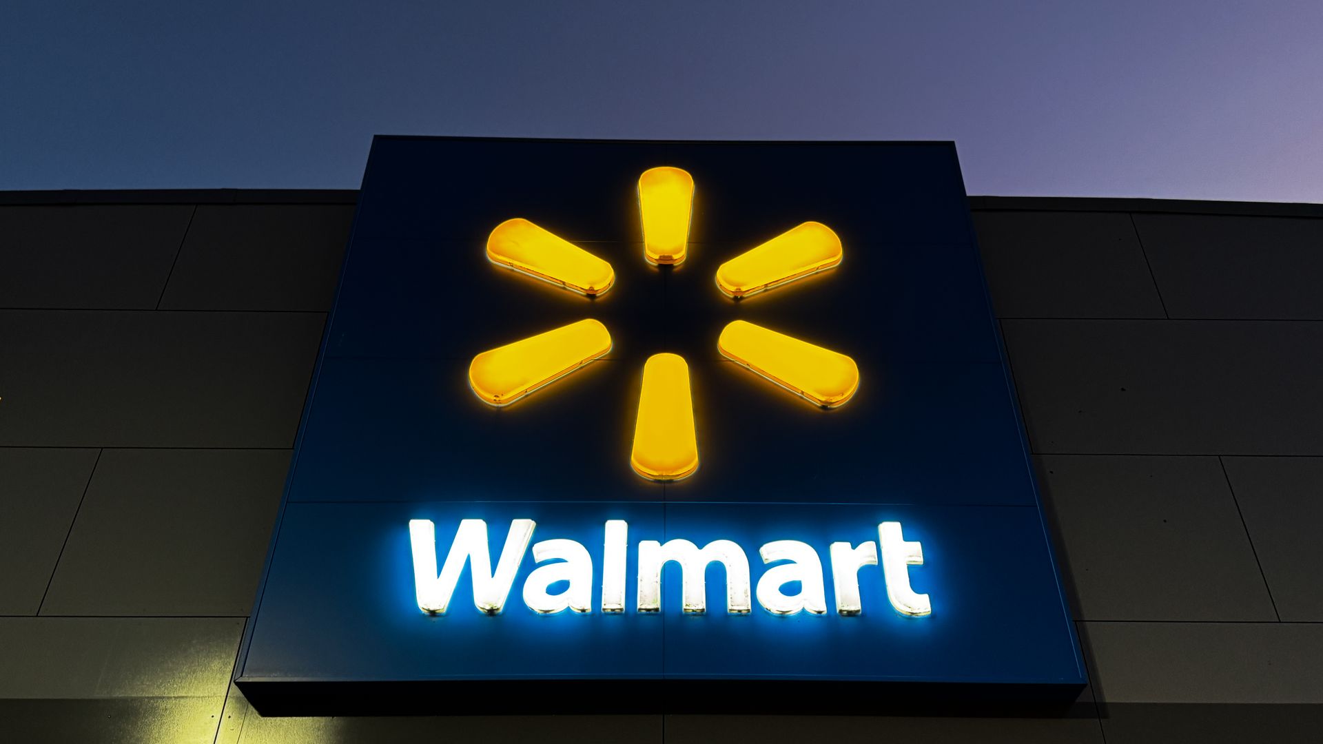 Illuminated Walmart logo with yellow spark symbol and white text on a blue square background against an evening sky.