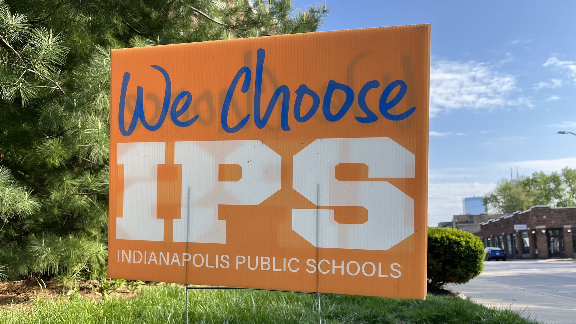 Orange Indianapolis Public Schools sign on a grassy verge reads "We Choose IPS" in blue cursive above large white IPS and the line "INDIANAPOLIS PUBLIC SCHOOLS". Trees, a street, and blue sky surround it.