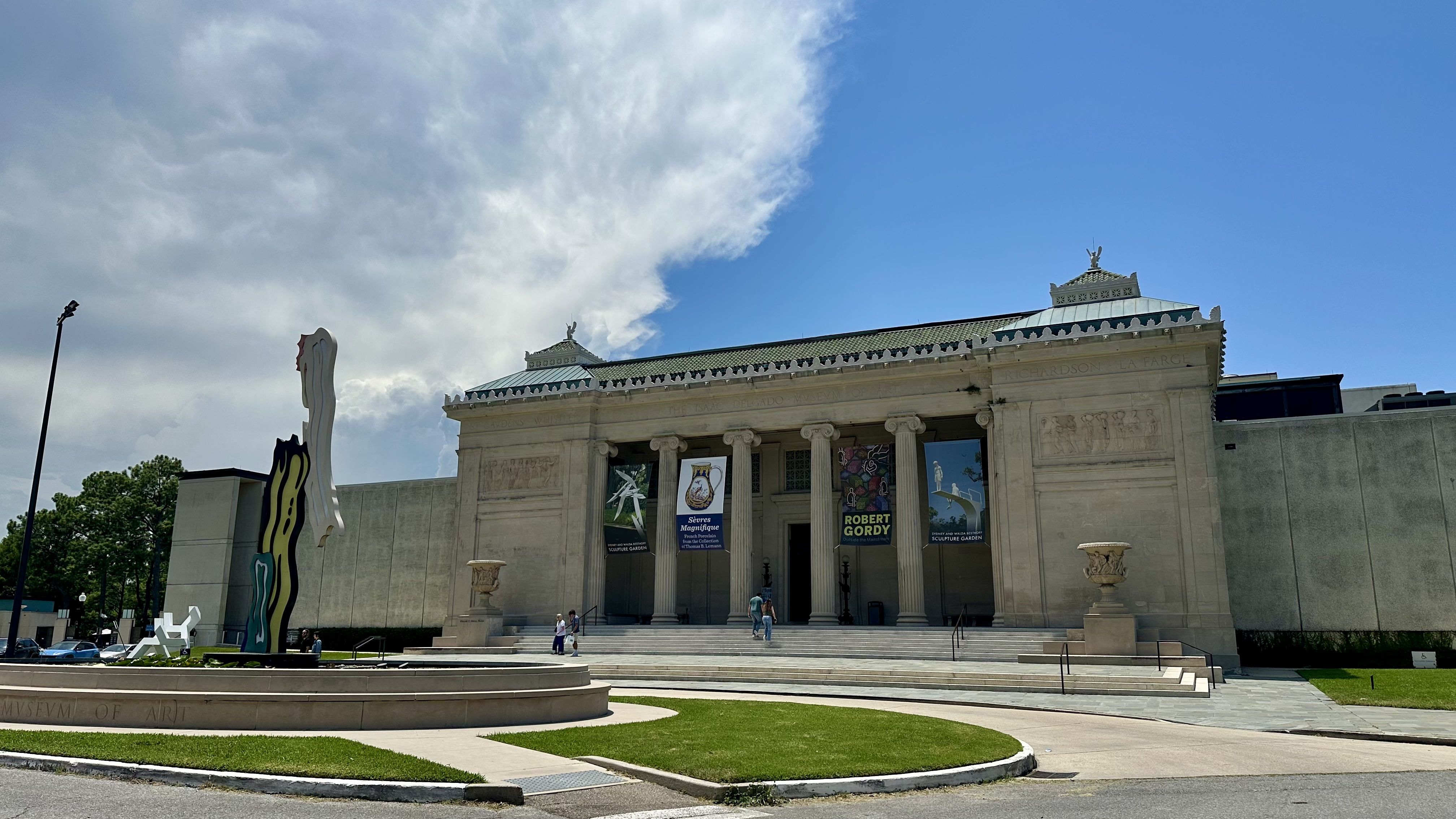 Neoclassical museum with tall columns, decorative frieze, and a green-tiled roof. Banners hang at the entrance; a modern sculpture stands to the left. People walk the steps under a blue, partly cloudy sky.