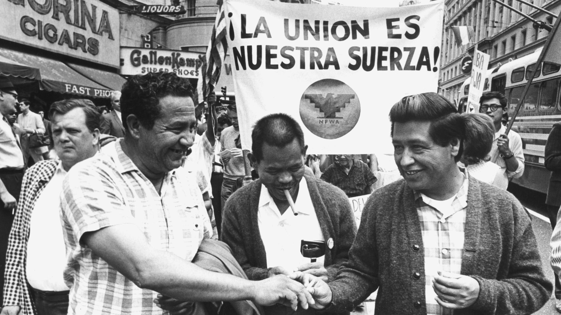 A black and white photo shows three men in the front of a protest line, all shaking hands. The man on the right is activist Cesar Chavez and in the middle is Larry Itliong, who is smoking a cigarette
