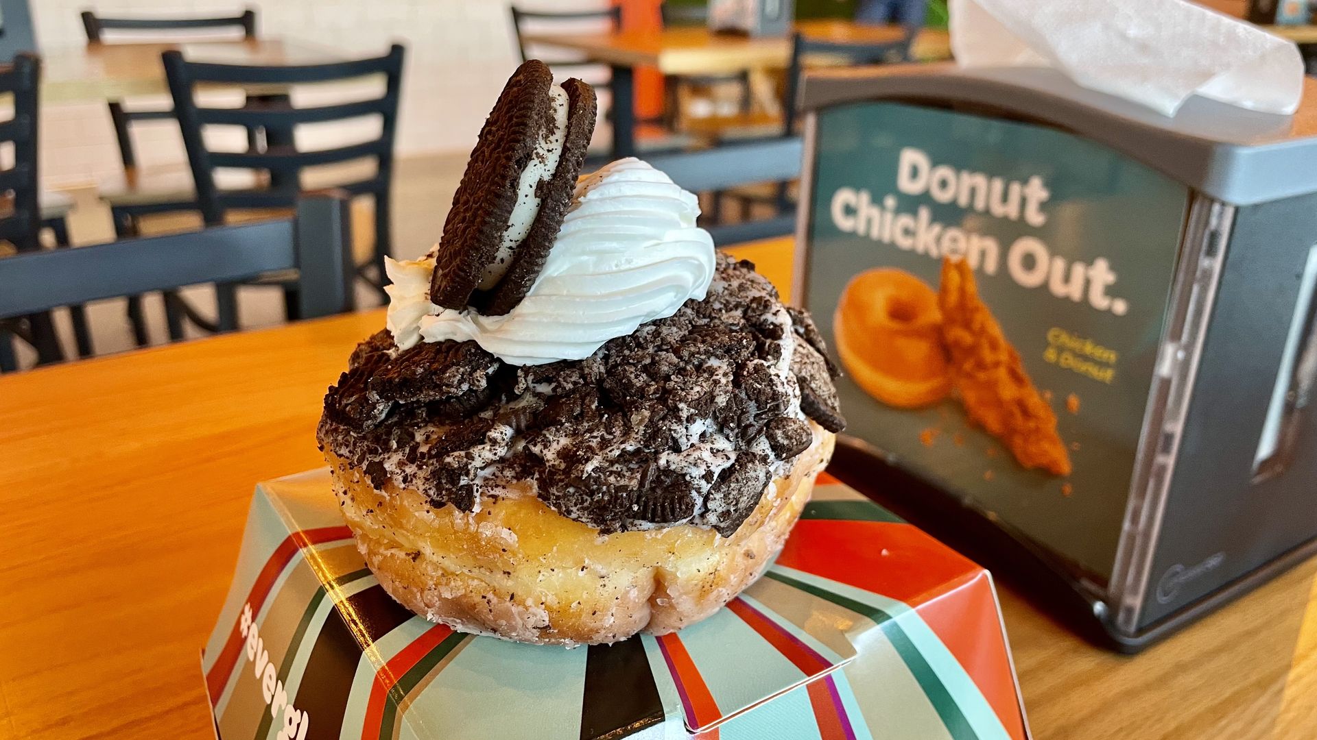 A donut topped with white cream, crushed chocolate cookies, and a whole chocolate sandwich cookie, placed on a colorful striped box on a wooden table with chairs in the background.