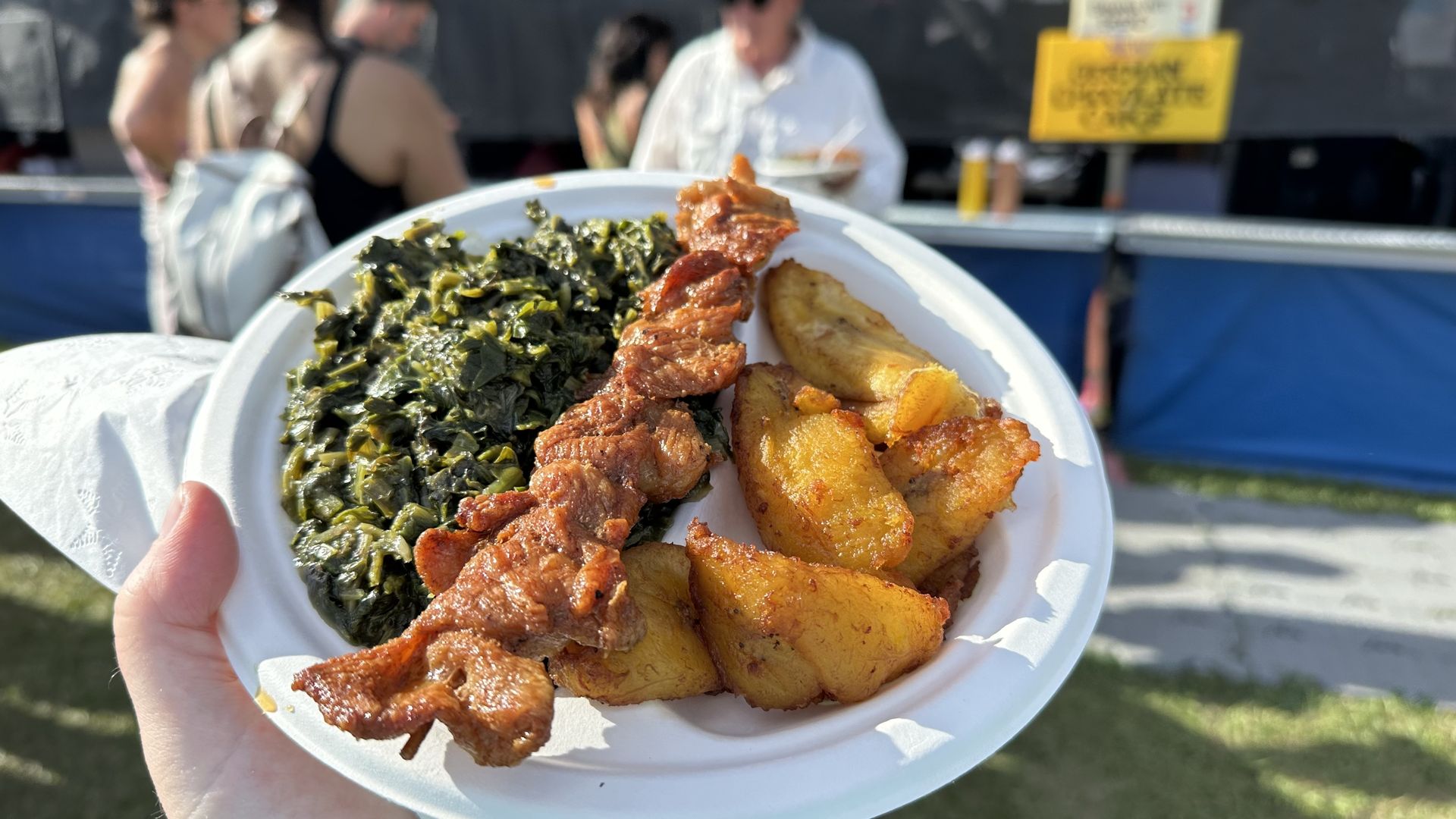 White disposable plate held outdoors with sautéed kale, crispy fried plaintains and sliced chicken. Blurry background shows people at an outdoor event and a yellow sign.