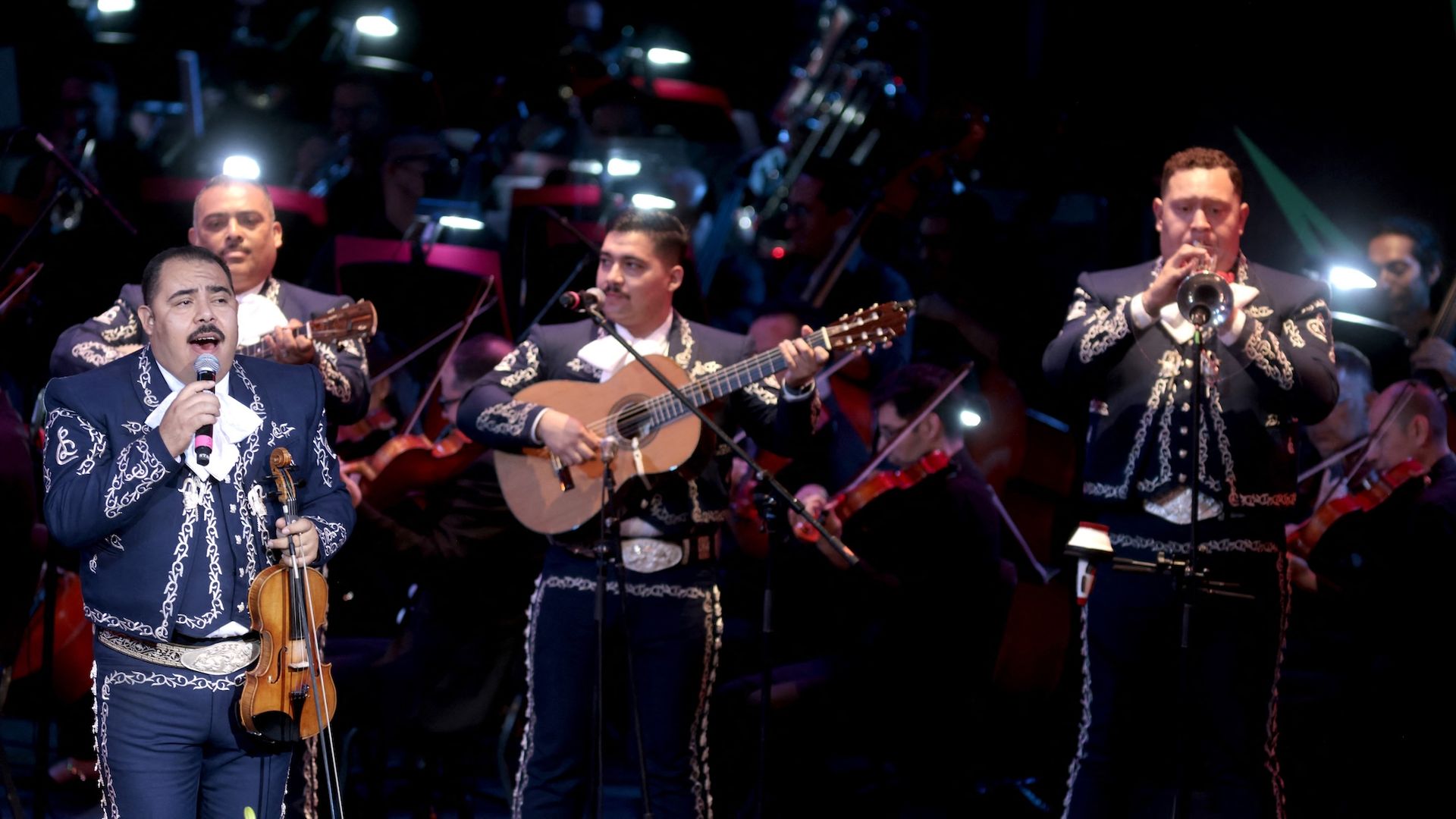 Musicians from the group Mariachi Los Camperos perform on stage in embroidered blue suits.