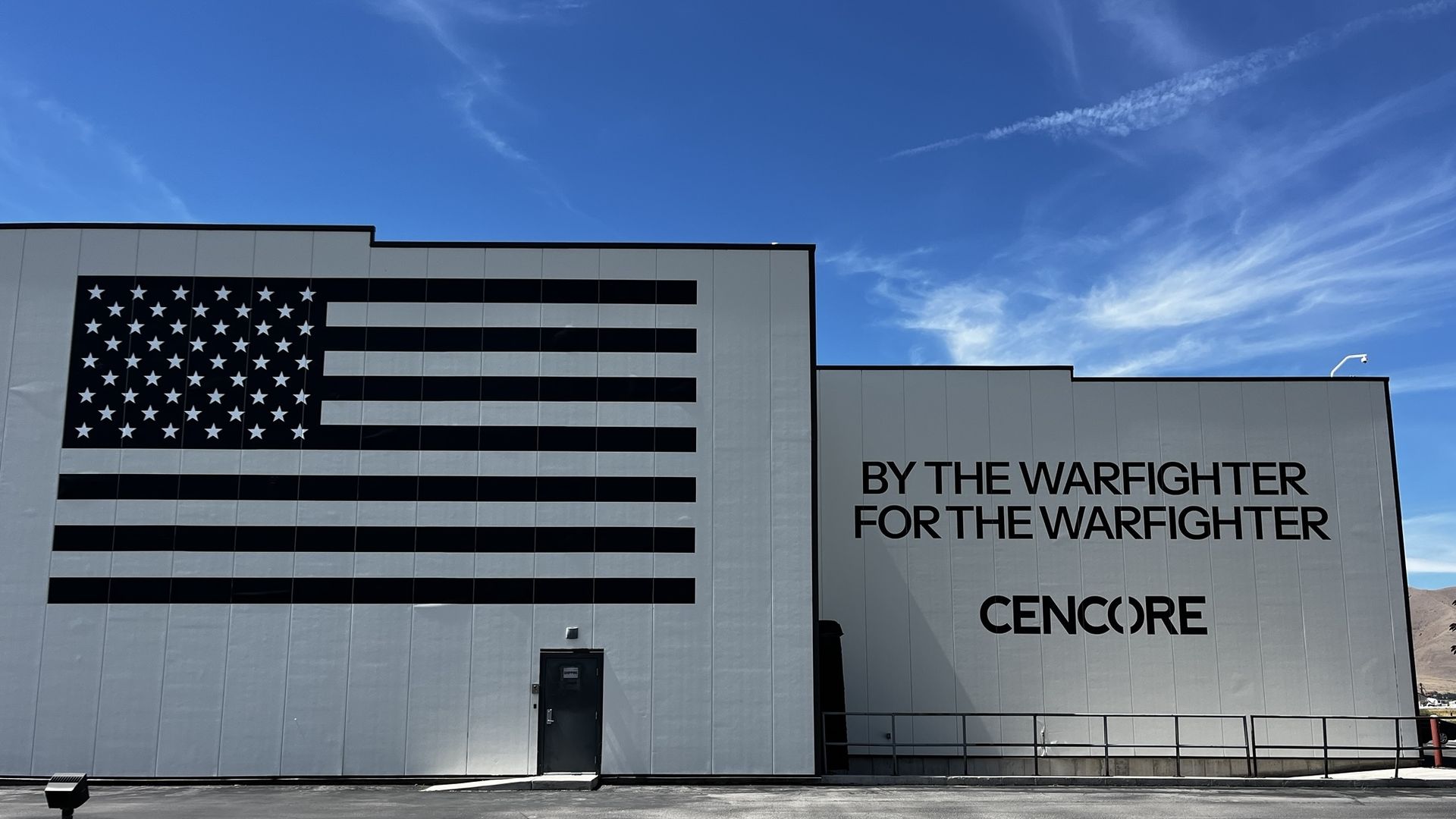 Exterior industrial building with a large black-and-white American flag mural on the left; on the right, bold black text reads "BY THE WARFIGHTER FOR THE WARFIGHTER".