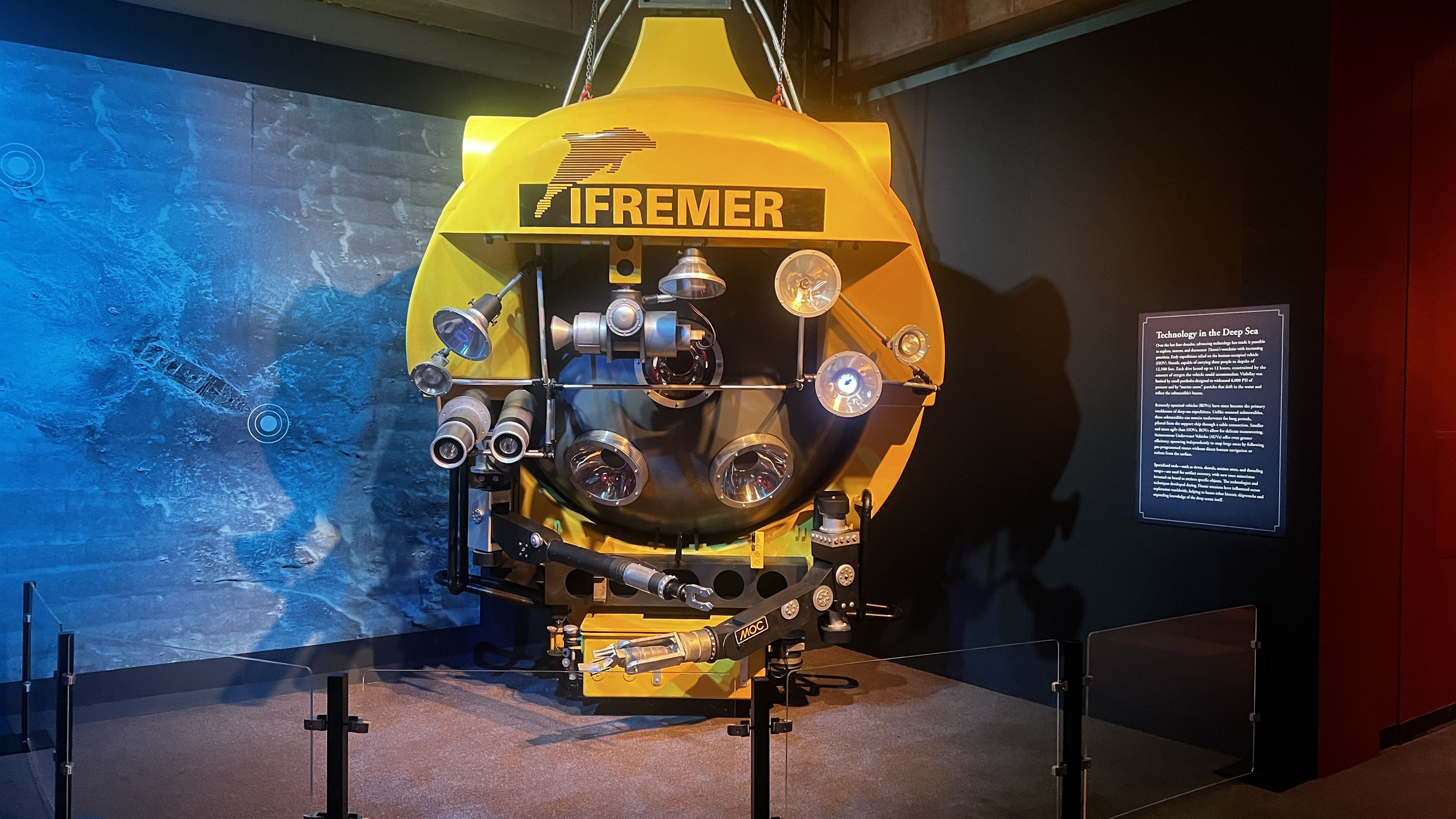 Yellow underwater research craft labeled IFREMER hanging from cables, with multiple lenses and robotic arms on a museum display; blue left wall and dark right wall surround it.