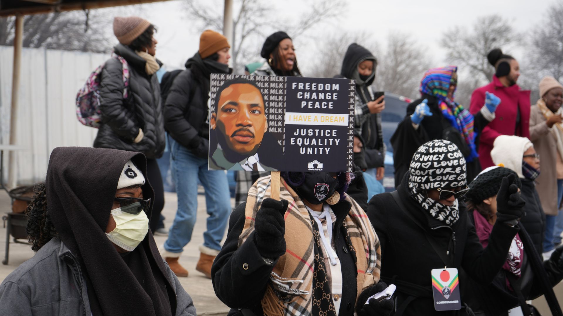 People dressed for cold weather at San Antonio's 2025 MLK March; one holds a sign with Martin Luther King Jr.'s portrait and the words: "Freedom, Change, Peace. I Have A Dream. Justice. Equality. Unity."