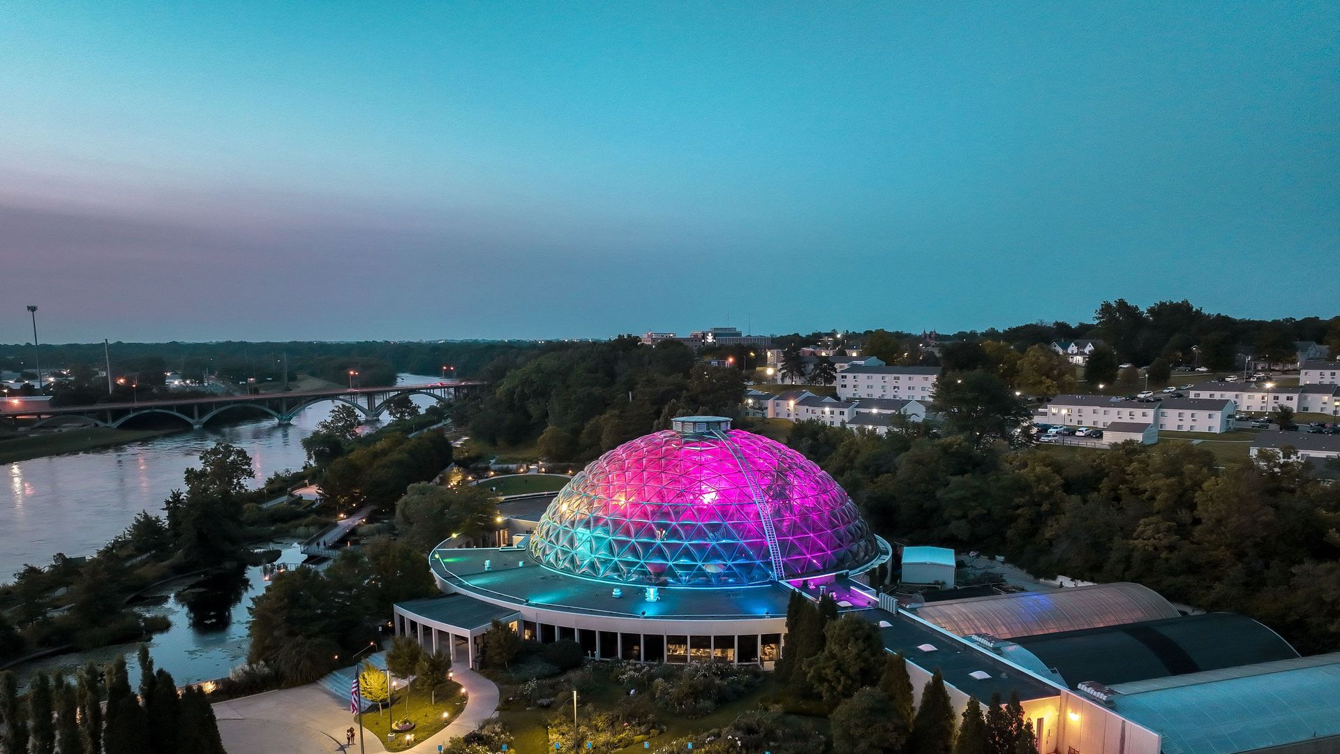 Aerial view at dusk: a colorful geodesic dome in the foreground, a river with a curved bridge on the left, and white apartment buildings lining the right, under a blue sky.
