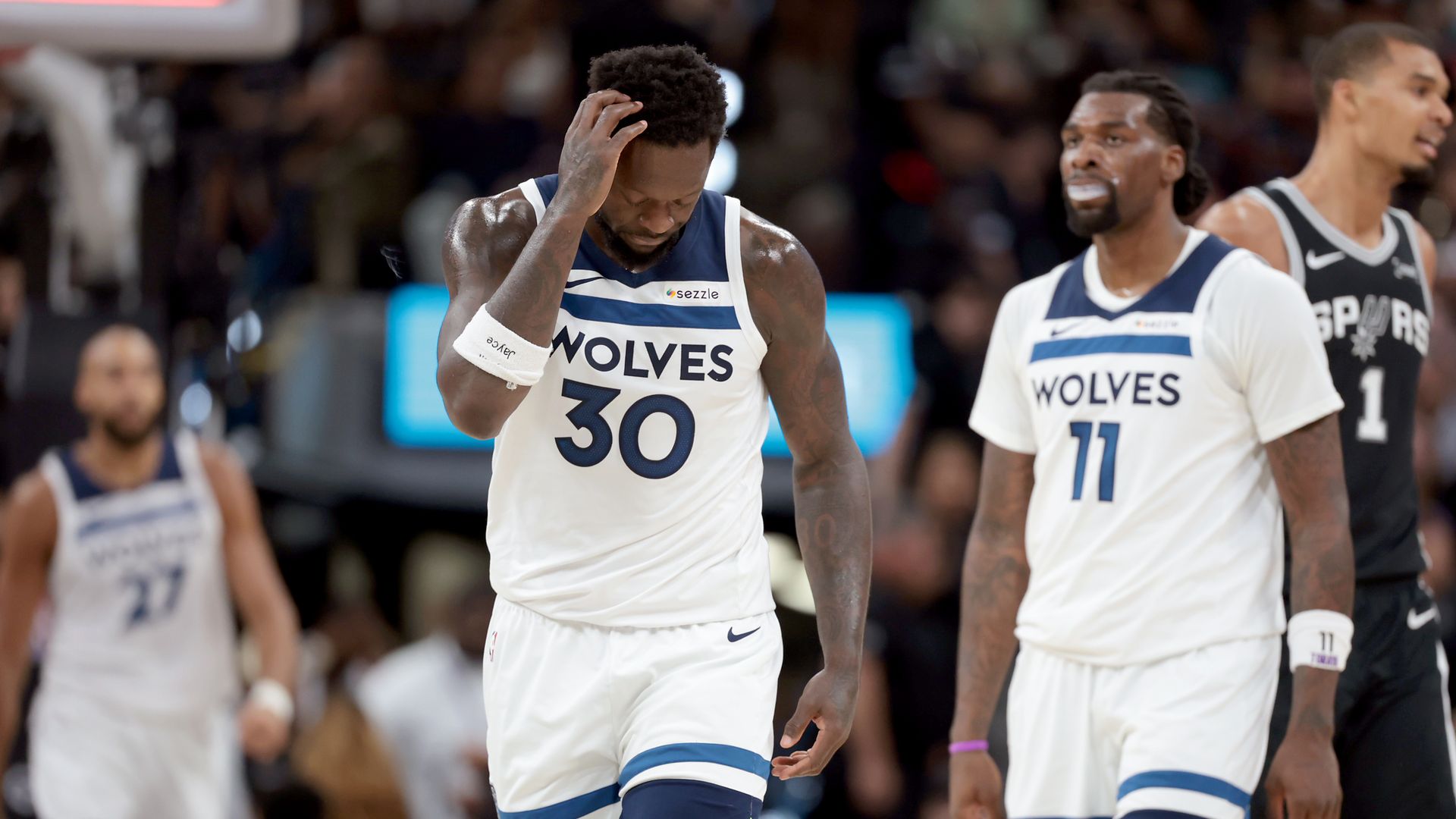 Two Minnesota Wolves in white jerseys with blue trim (#30, #11 beside him) walk on court as the center player wipes sweat from his forehead; a Spurs player in black is blurred in background.