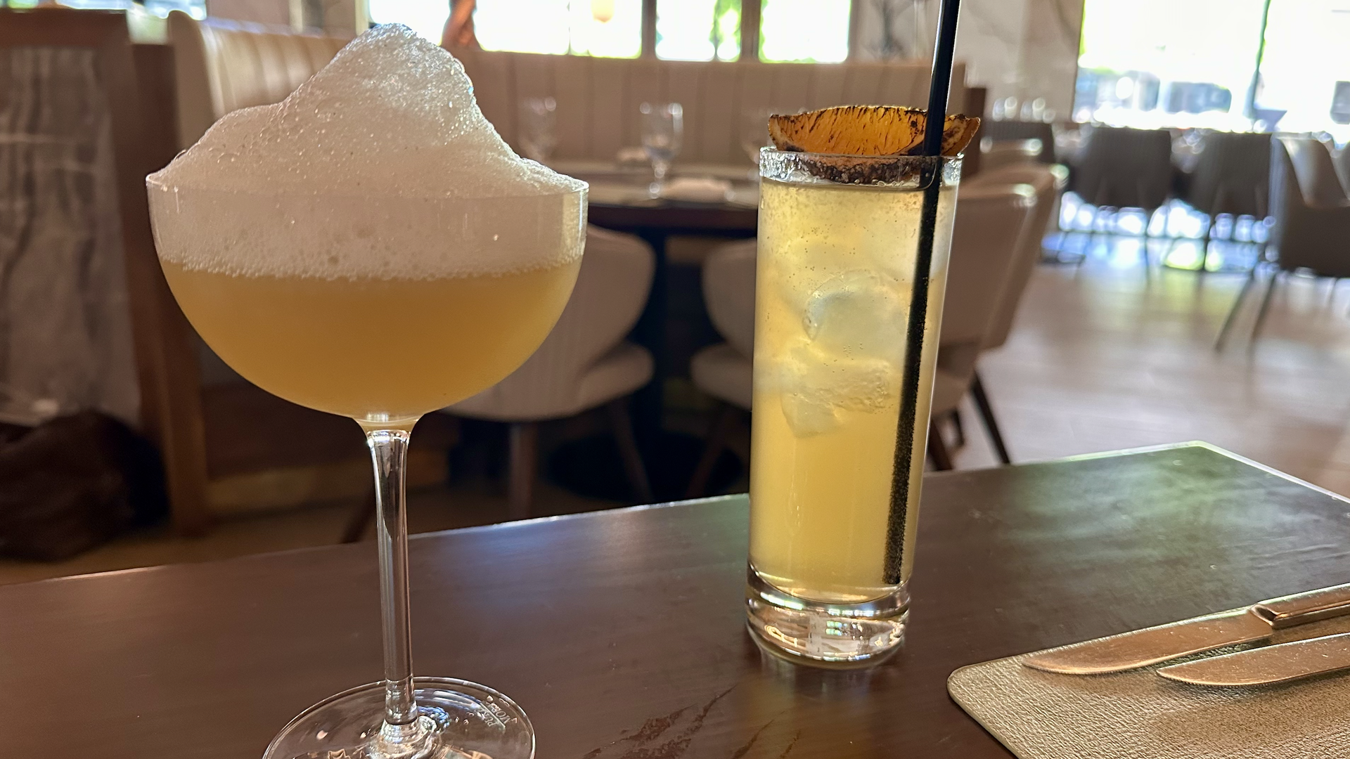 Two cocktails on a wooden table in a bright restaurant: a frothy pale yellow drink in a coupe on the left; a tall iced drink with a salted rim and orange slice on the right.