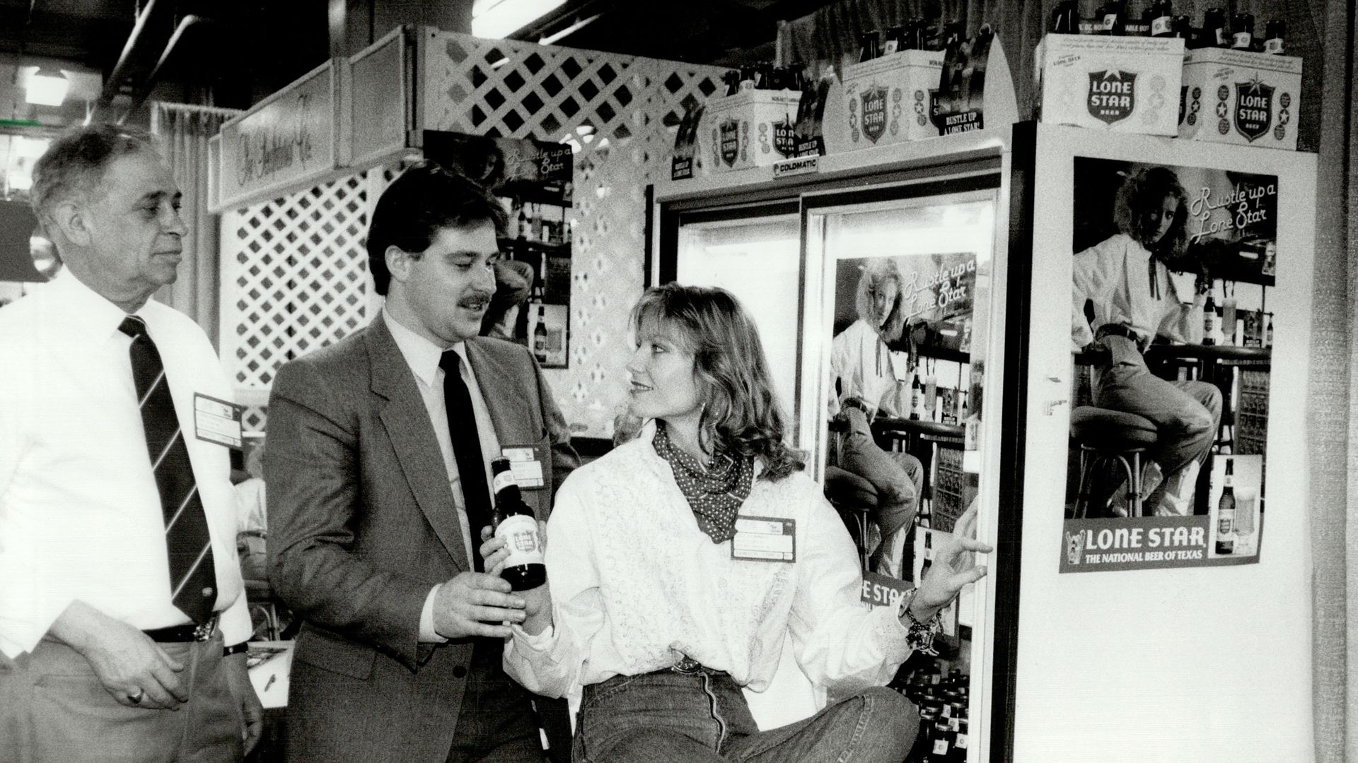 Miss Lone Star Beer Lisa Downey, pictured here in this black and white photo, helped introduce the San Antonio beer to Ontario in 1986. She is seen handing a Lone Star to a man in a suit with Lone Star posters around them.