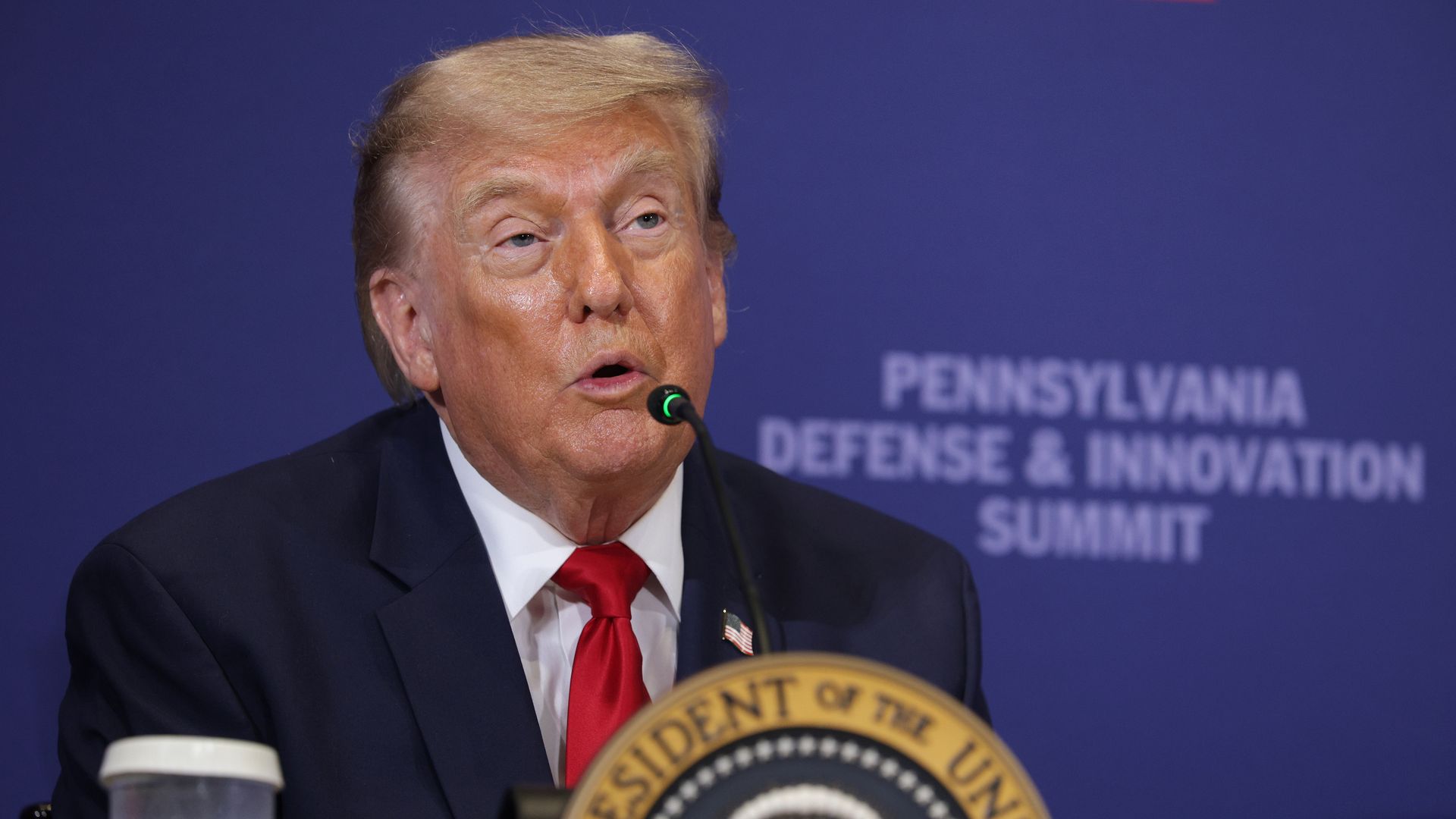 President Trump in a dark suit and red tie speaks into a microphone at a podium with the presidential seal, against a blue backdrop reading "Pennsylvania Defense & Innovation Summit".