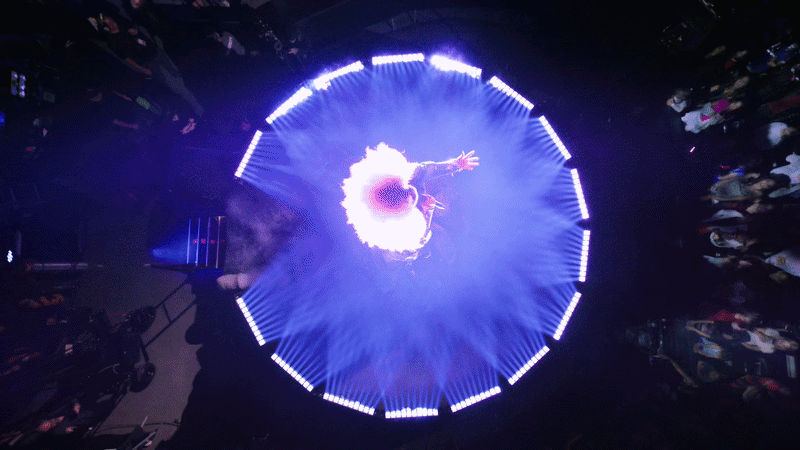 A performer stands in the center of a circular LED ring on a top-down stage, arms outstretched, with a bright purple-white halo and blue haze; crowd surrounds the dark venue.