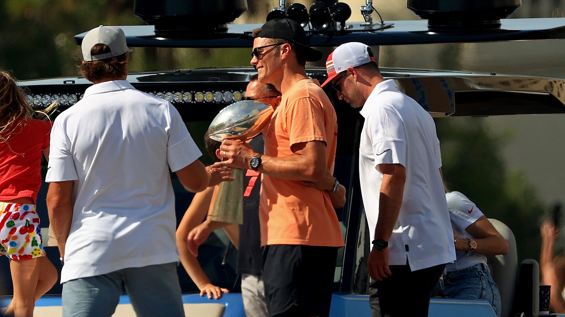 Group of people on a boat, one man in an orange shirt holding a silver trophy resembling a football, others wearing white shirts and caps, sunny outdoor setting.