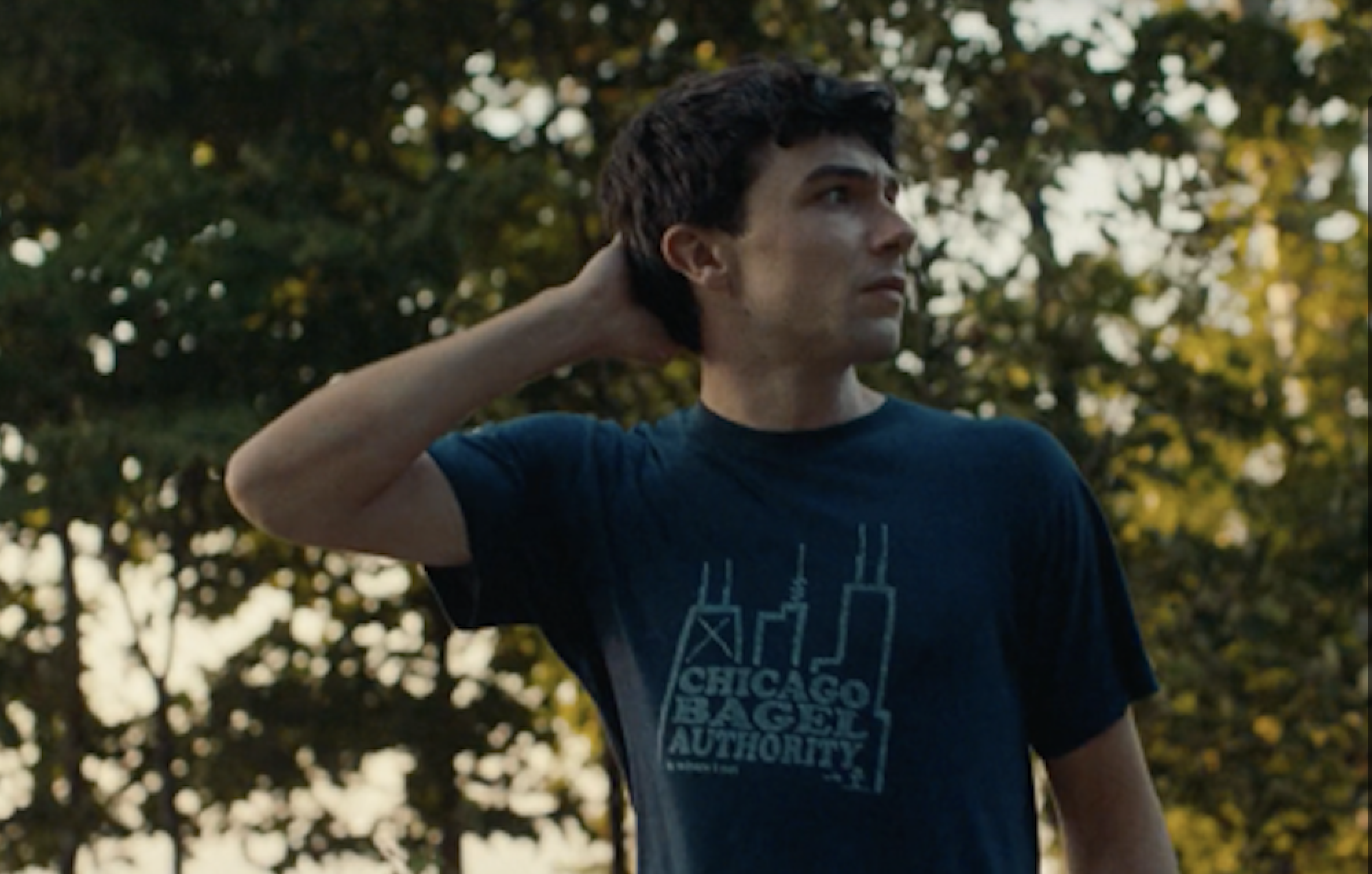 Young man with dark hair outdoors among green trees, looking to the right, his right hand resting behind his neck, wearing a blue T-shirt with a graphic that reads CHICAGO BAGEL AUTHORITY.