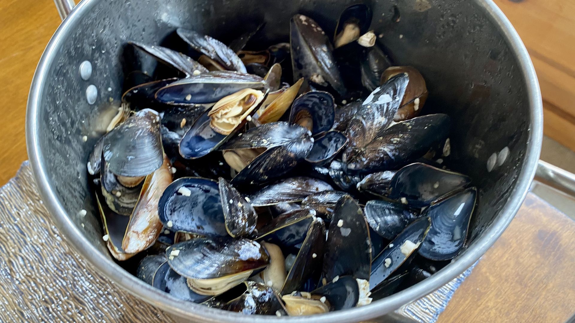 A stainless steel pot filled with mussels on a wooden table with a silver table runner. 