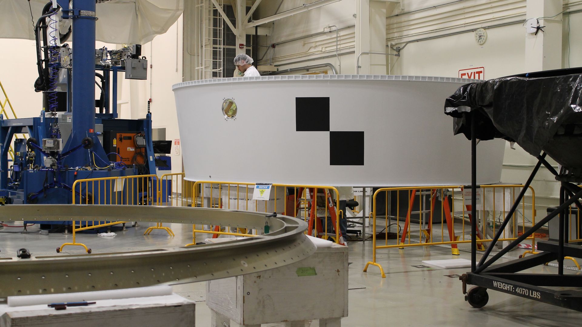White circular structure with black squares inside a large industrial space, safety barriers around it and a person in protective gear nearby, various machinery and equipment visible.