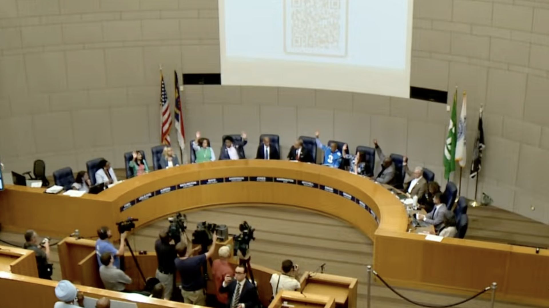 Wide view of a large council chamber with a curved wooden dais and blue seats. Officials sit around, many raising their hands. Flags line the back wall as cameras and reporters fill the foreground.
