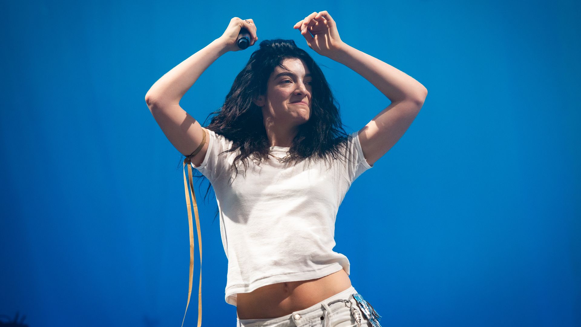 Person with long dark hair wearing a white cropped t-shirt and white pants, holding a microphone with a gold ribbon, performing against a solid blue background.