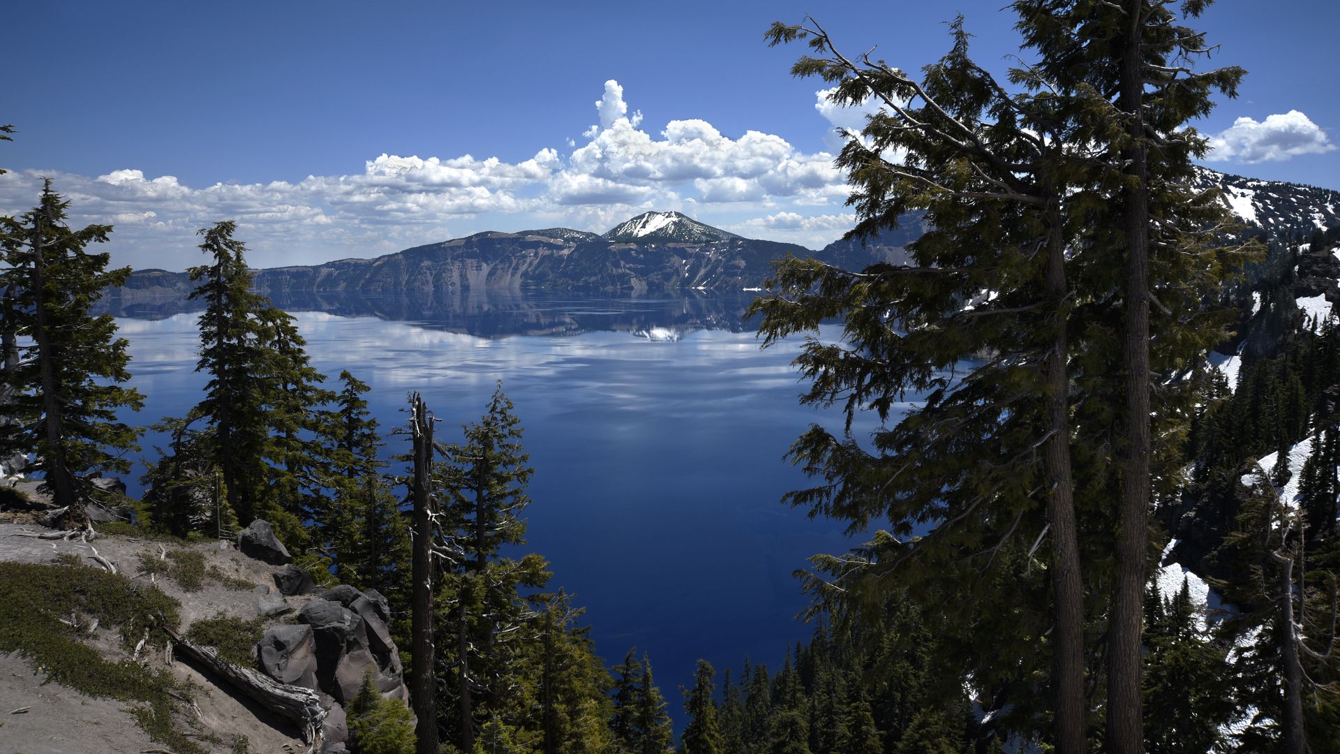 A deep blue lake framed by tall evergreen trees, snow-capped mountains in the distance, and a rocky foreground; a bright sky with white clouds reflects on the calm water.