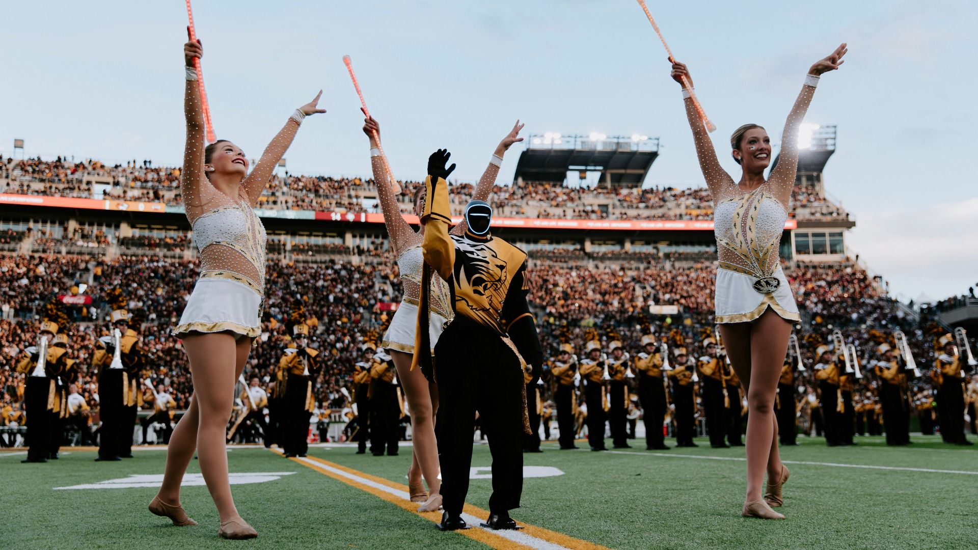 Three baton twirlers in sparkling white-gold costumes raise pink light batons on a football field, flanked by a central masked performer; a large crowd in a stadium under a blue sky.