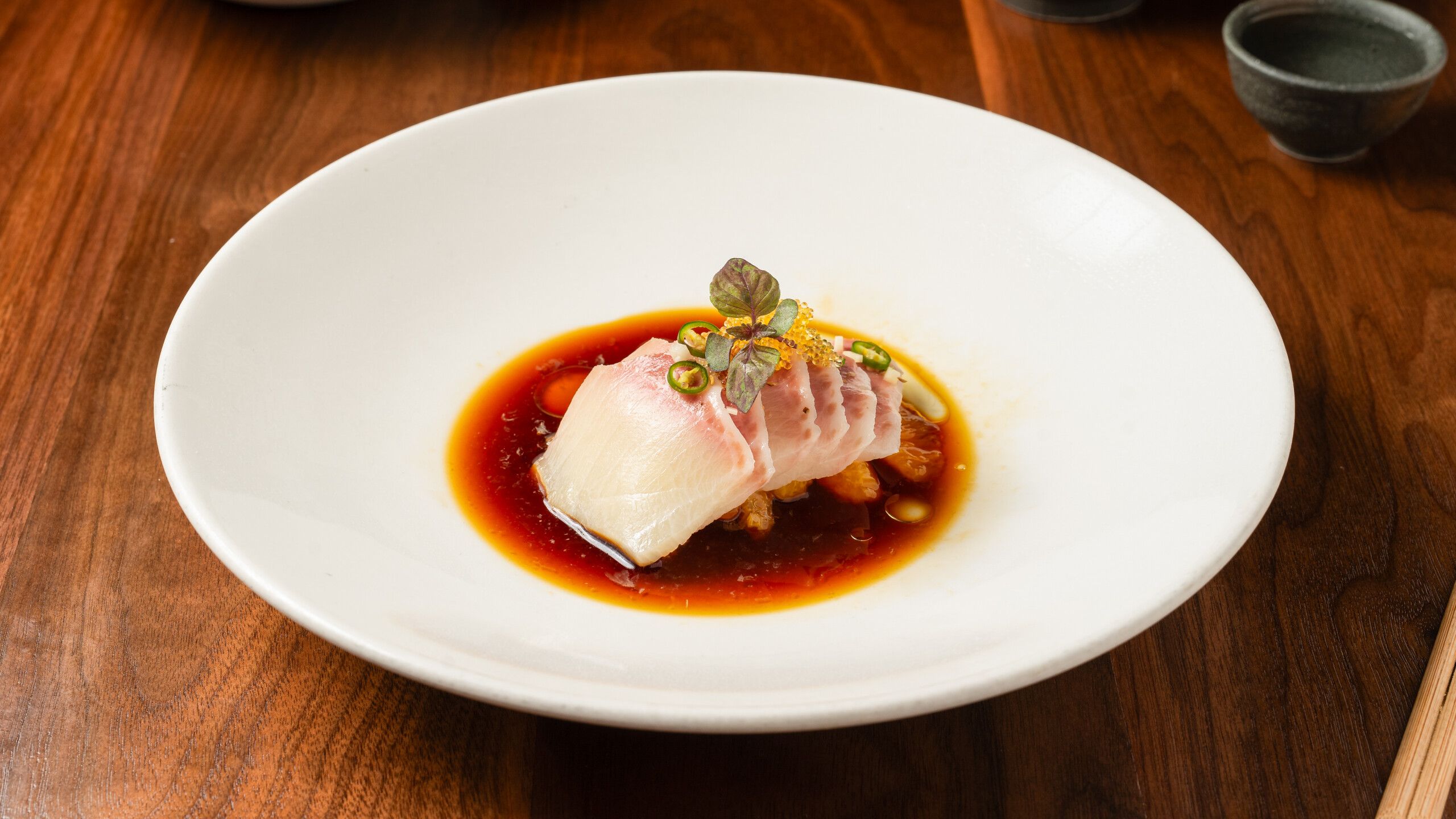 Sliced white fish sashimi arranged on a red sauce, garnished with microgreens, green chili slices, and a yellow crunchy topping on a white plate set on a wooden table.