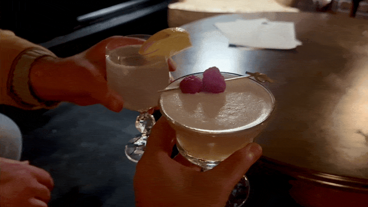 Two hands holding cocktails in elegant glasses, one garnished with a lemon slice, the other with two raspberries on a cocktail pick, against a dimly lit bar table background.