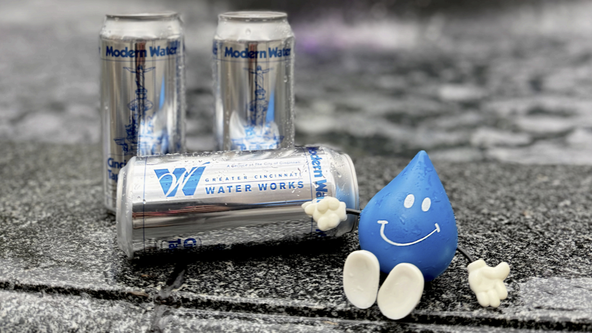 Three aluminum water cans labeled Greater Cincinnati Water Works lie on a wet stone surface beside a blue, smiling water drop figure wearing white gloves and shoes.