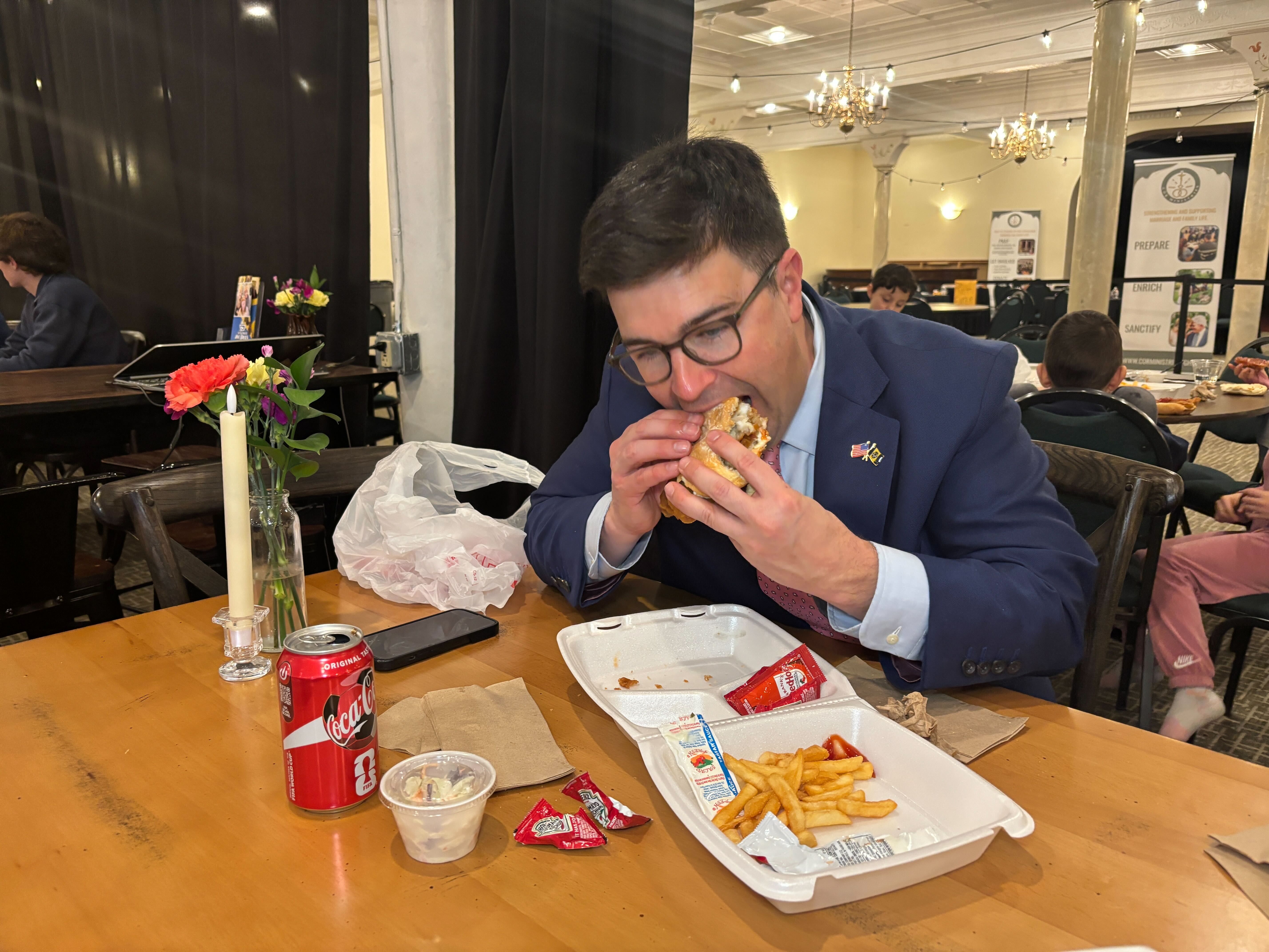 Man in a blue suit and glasses eating a sandwich at a wooden table with a takeout container of fries, a can of Coca-Cola, a small salad cup, and condiment packets, in a decorated indoor venue.