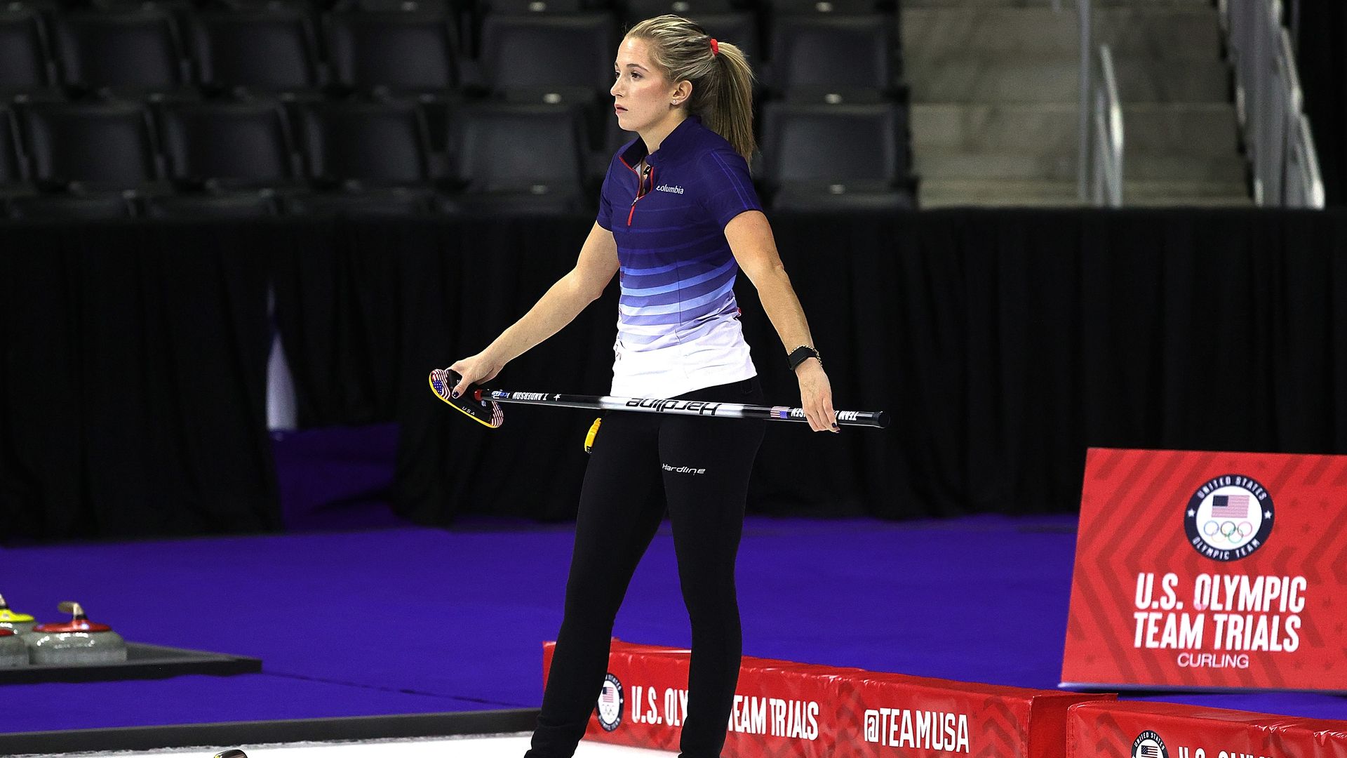 Female curler in purple and white shirt stands on ice holding a curling broom, next to a curling stone. Background shows U.S. Olympic Team Trials curling signage in red and blue.