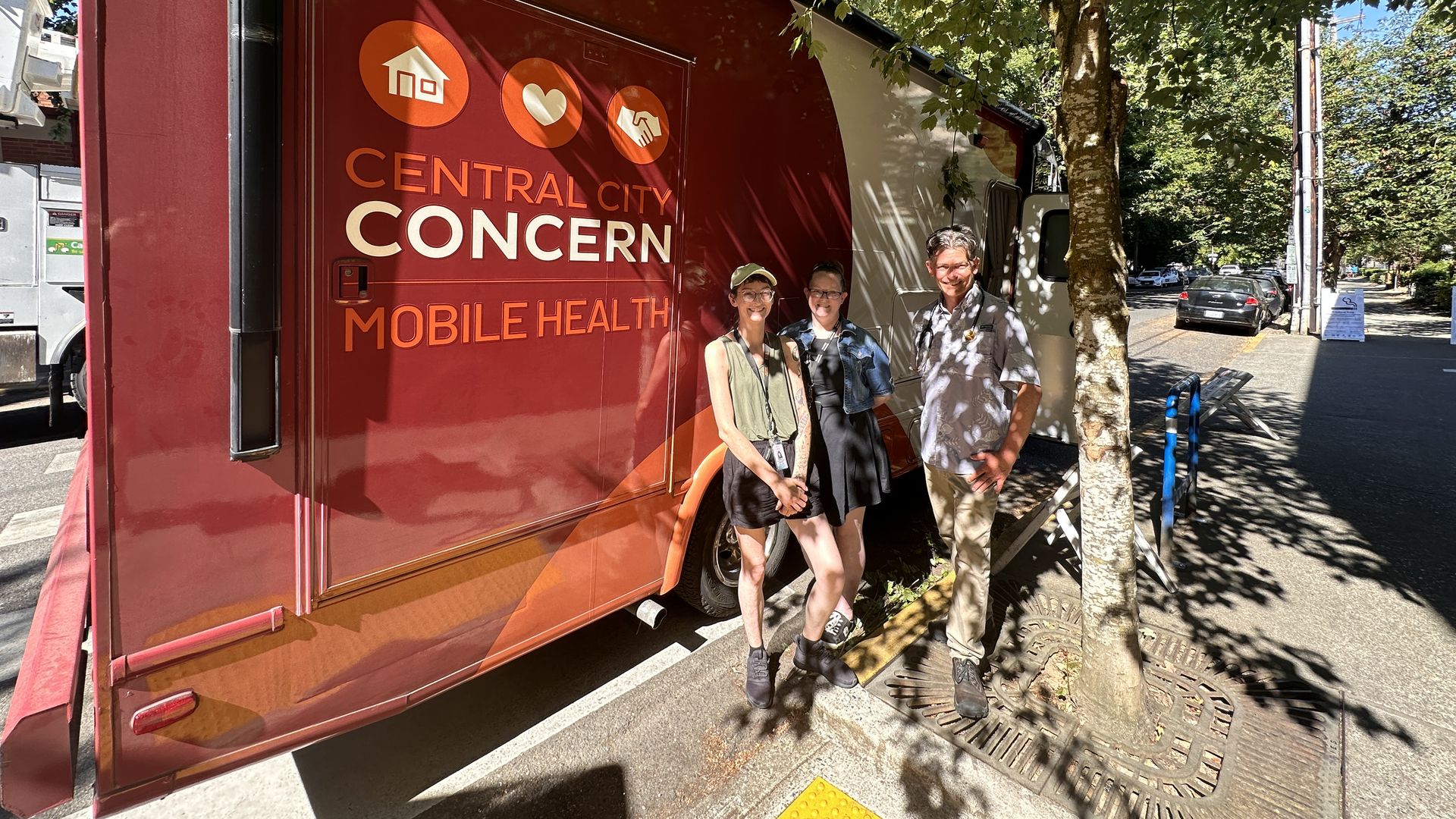 Three people in dappled sunshine standing beside a mobile medical unit marked Central City Concern.