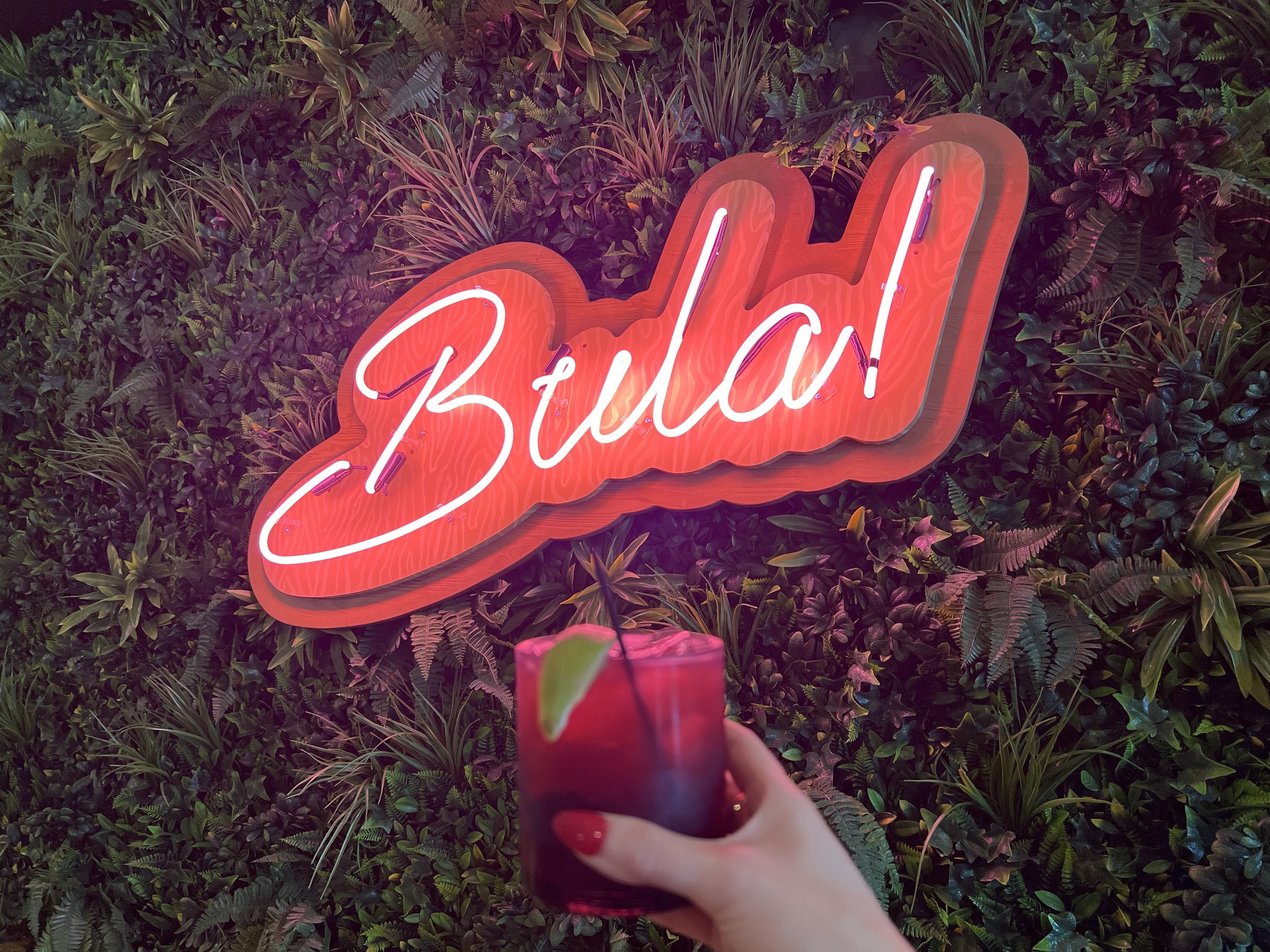 A hand holding up a cocktail in front of a plant wall that has "bula!" as a  neon sign