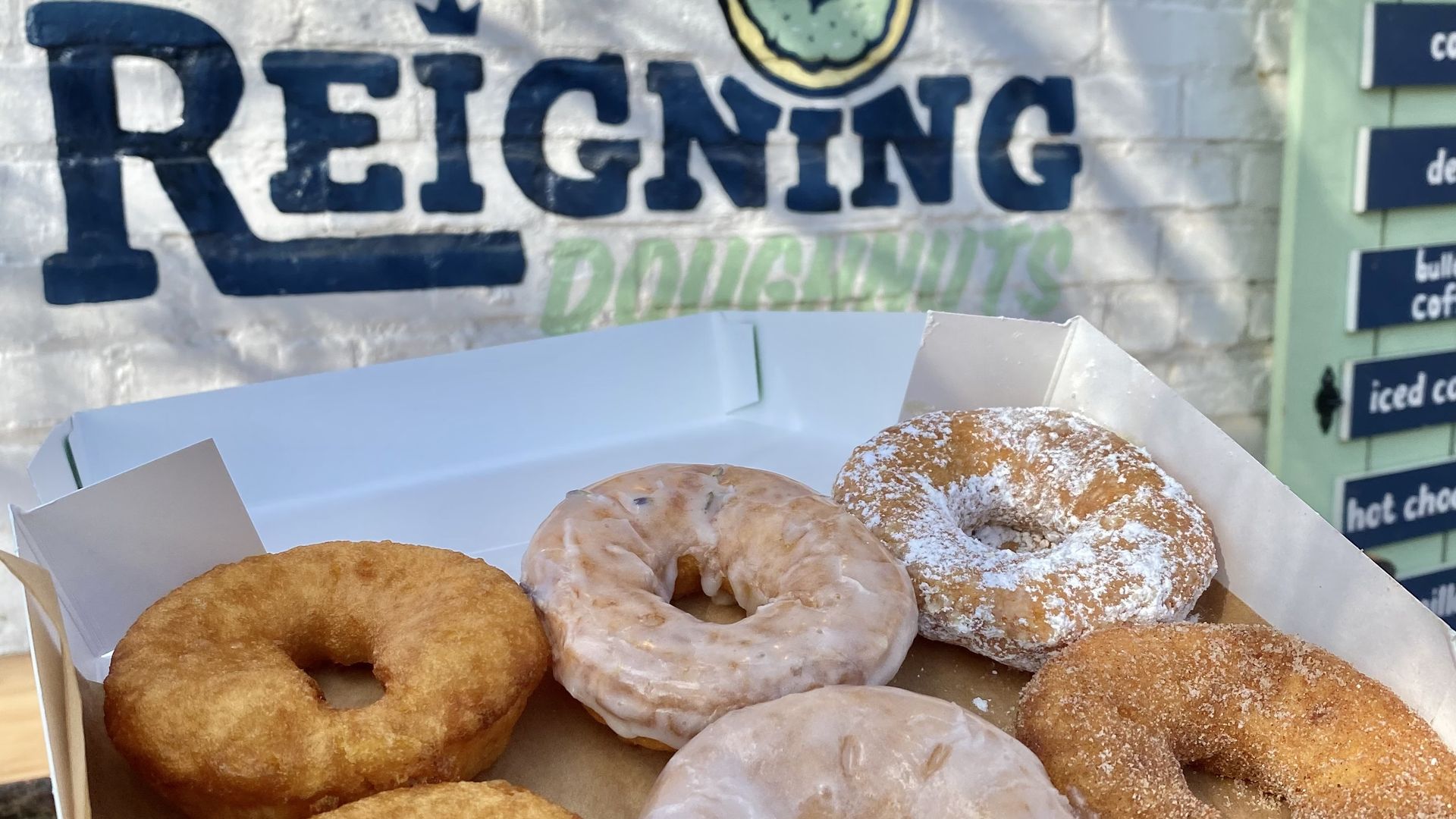 Box of six assorted doughnuts including glazed, powdered sugar, and cinnamon sugar, held in front of a brick wall with a painted sign for Reigning Doughnuts.