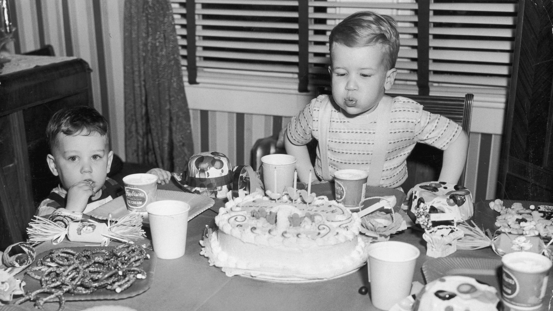 Black and white picture of a child blowing his birthday candles surrounded by cookies, candy and other sweets and snacks while another boy looks at the camera