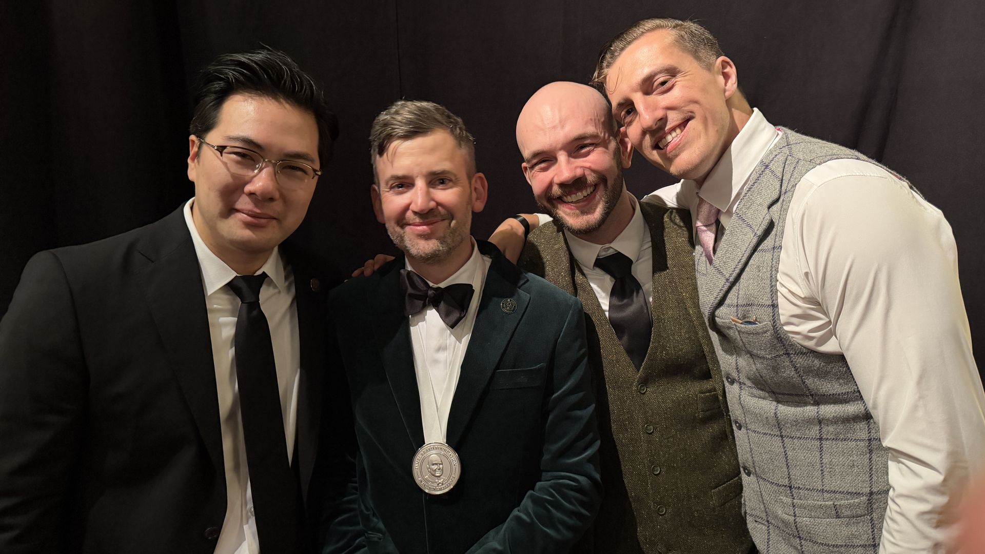 Left to right: a man in a black suit and glasses; a man in a velvet suit with a bow tie; a bearded man with a medal; and a man in a gray plaid vest.