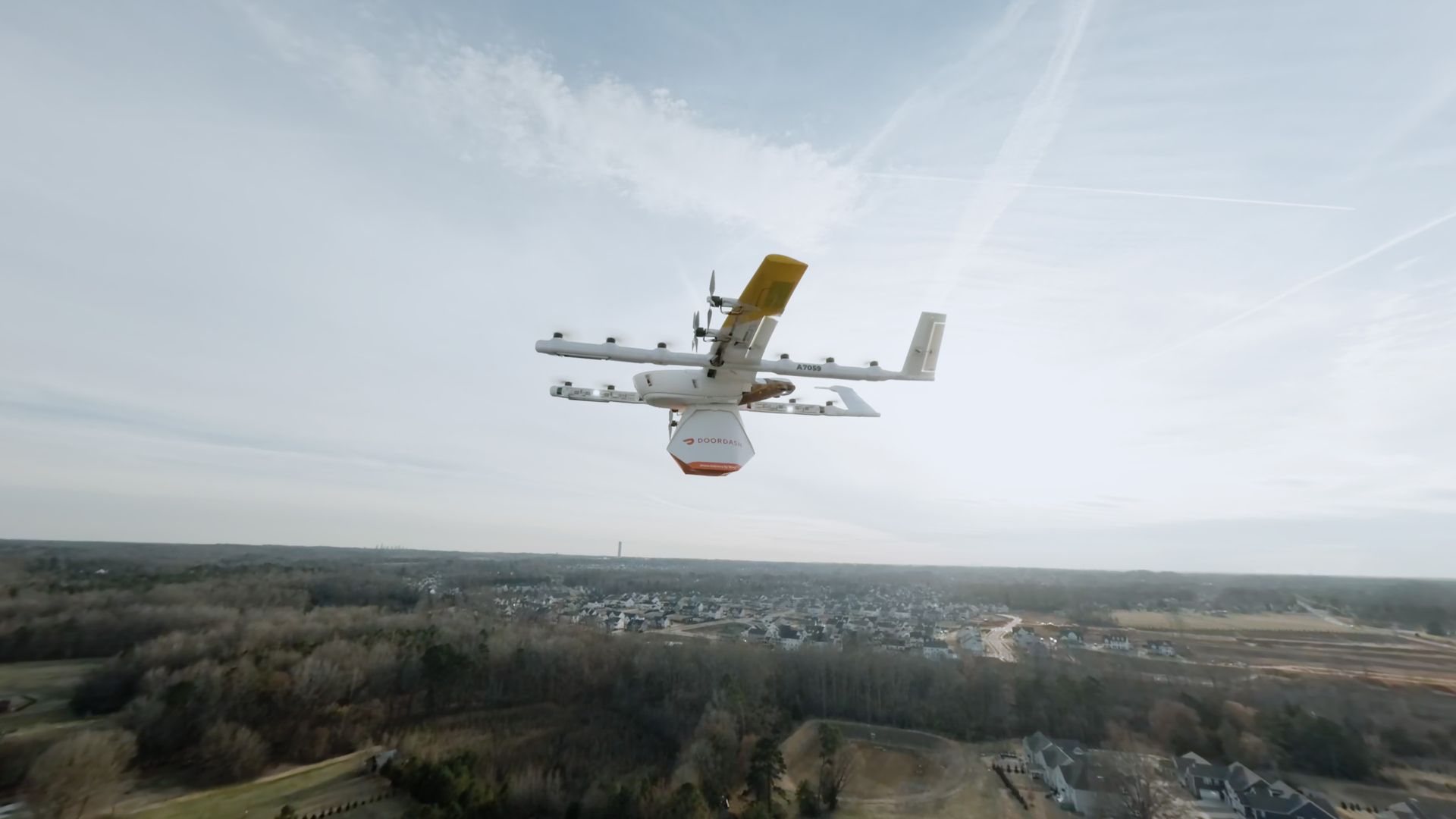 White delivery drone with a yellow wingtip flies over a suburban neighborhood, carrying an orange hexagonal Doordash payload pod beneath its belly against a pale blue sky.