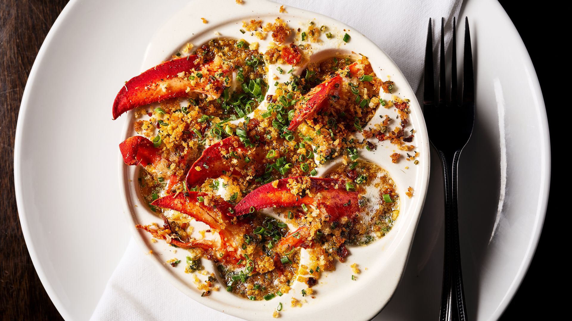 Creamy lobster bake in a white ramekin topped with golden breadcrumbs and chopped chives; red lobster tails visible, set on a white plate with a napkin and silverware on a dark wooden table.