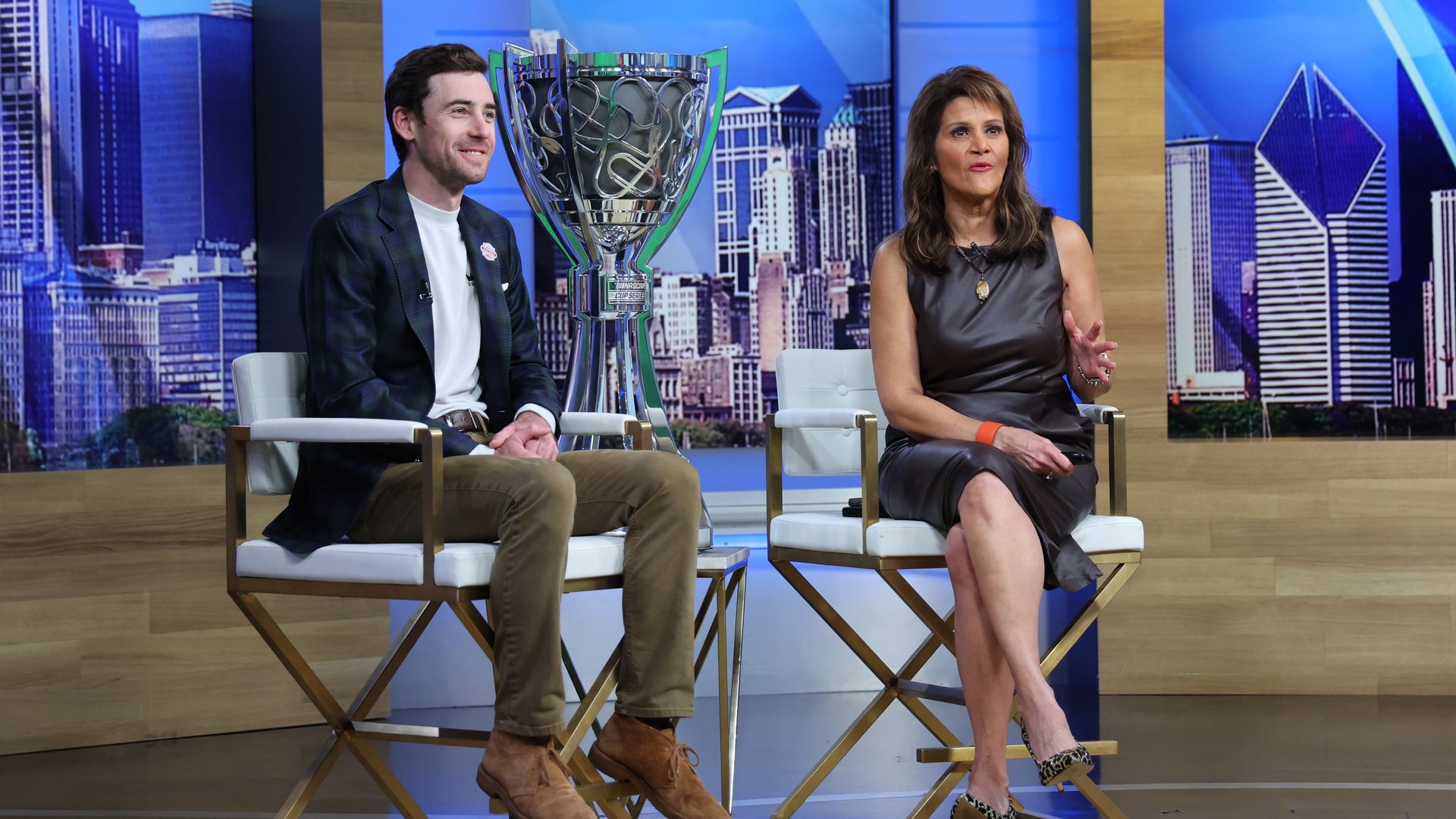 Two hosts sit on white-and-gold chairs on a TV studio set, smiling, with a large silver trophy framed between them. A city skyline backdrop and wood panels surround the scene.