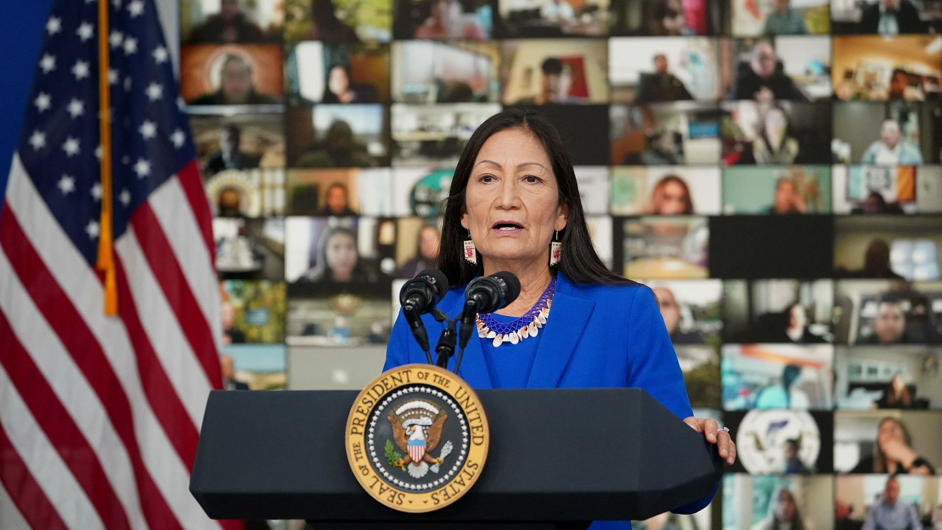 Photo of Deb Haaland speaking from a podium with a mosaic of Zoom video boxes showing different faces behind her