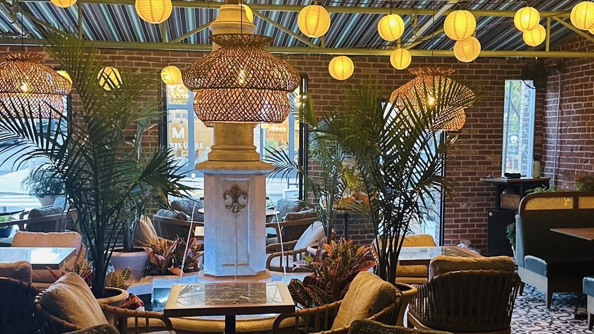 Cozy indoor café with large woven lamps, yellow paper lanterns, brick walls, lush green plants, and comfortable cushioned chairs around small tables.