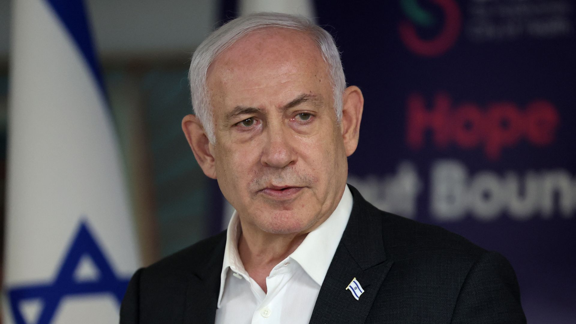 Israeli Prime Minister Benjamin Netanyahu, wearing a dark gray suit, white shirt and standing in front of an Israeli flag.