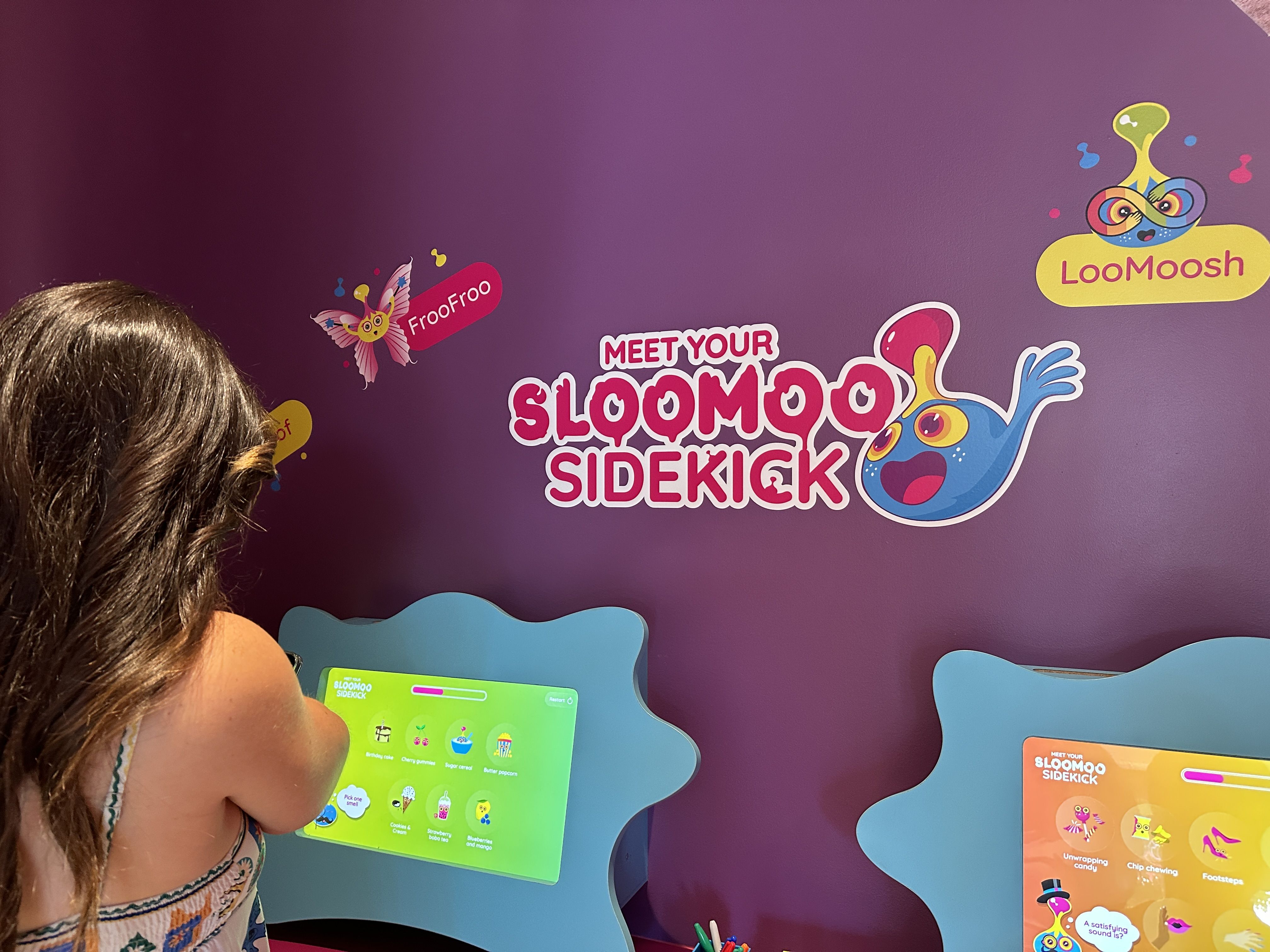 Purple wall with bright cartoon slime characters and logos: "Meet Your Sloomoo Sidekicks", "FrooFroo", and "LooMoosh". A child with brown hair looks at a bright green tablet.