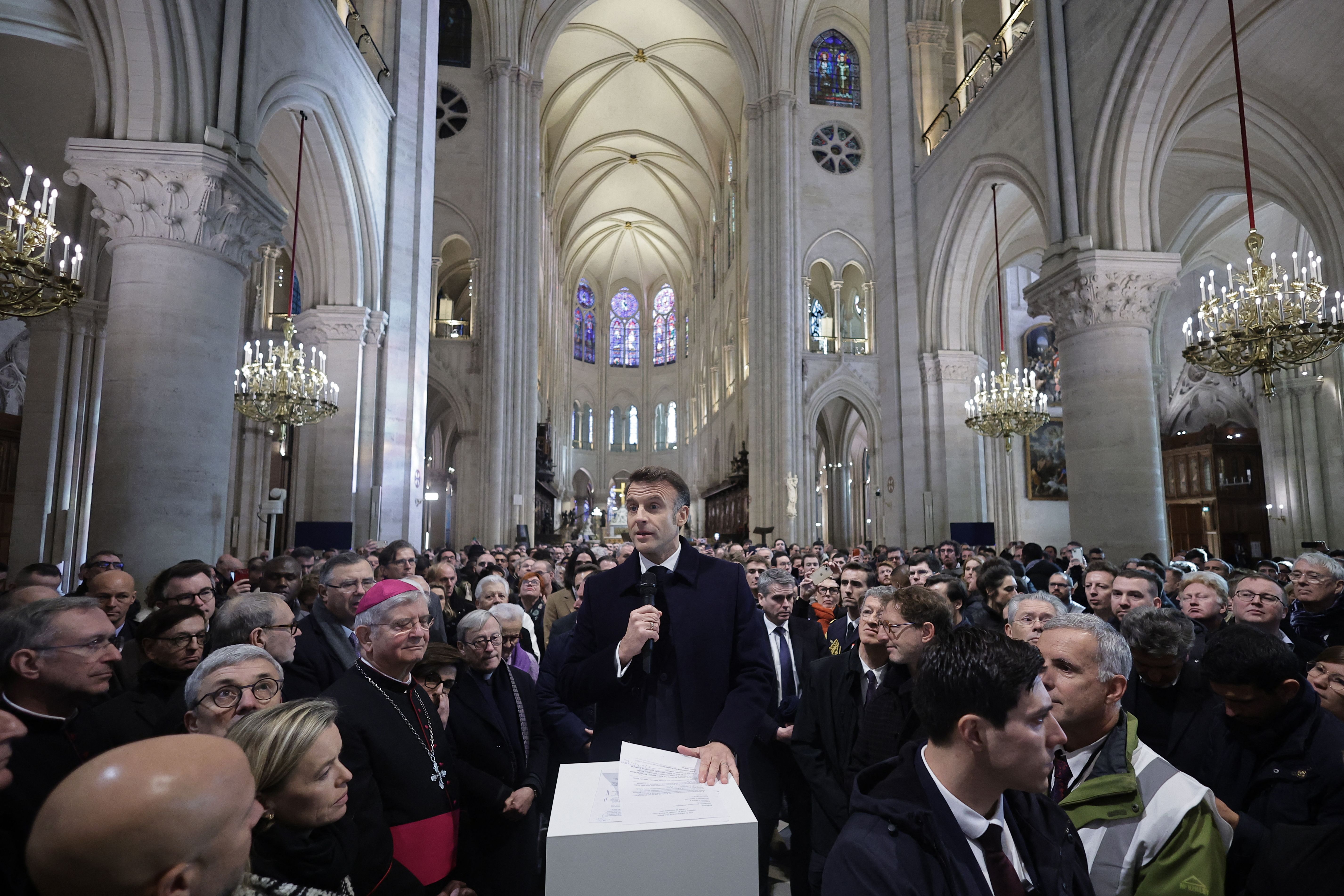 Macron speaks to a large crowd within the Notre Dame. He holds a microphone. 